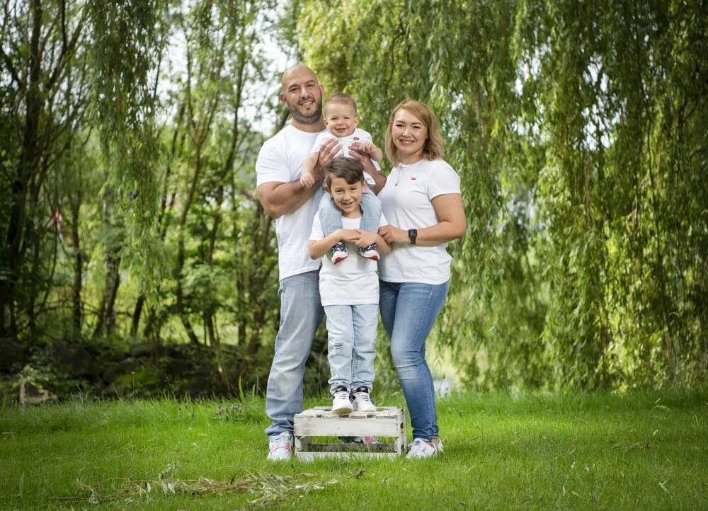 Outdoor Familienfotoshooting: Eltern mit zwei Söhnen auf einer Holzkiste unter Trauerweiden