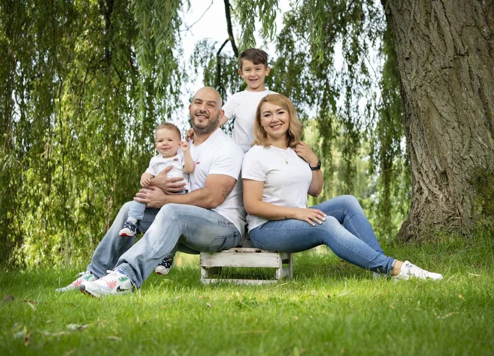 Familie in weißen Shirts sitzt entspannt auf einer Holzbank im Gras vor einem großen Baum