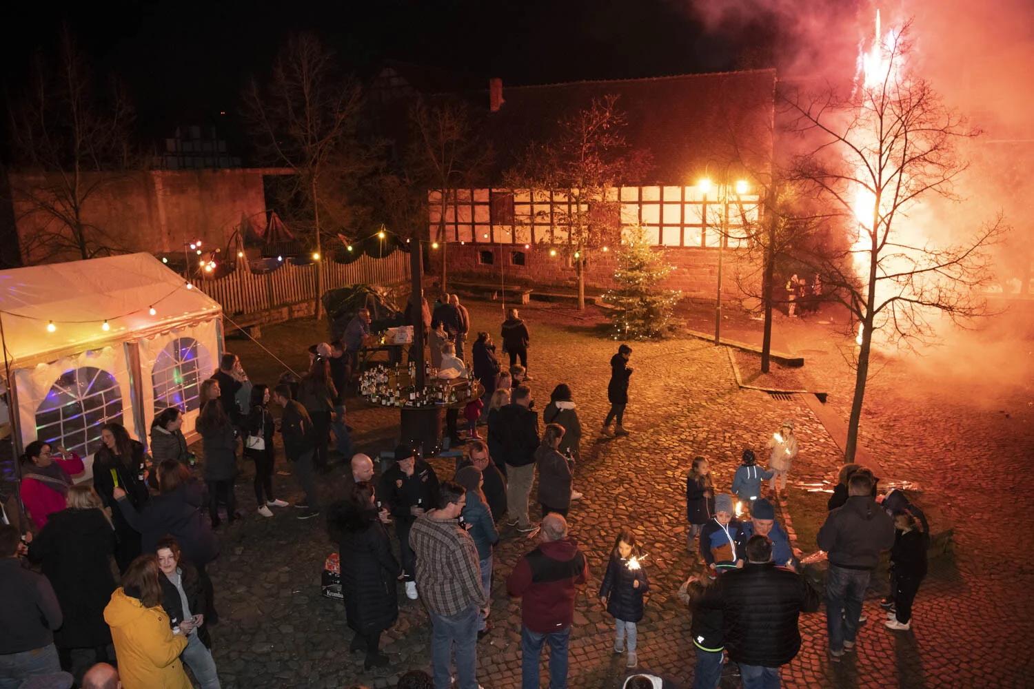 Silvesterfeier in Speckswinkel: rotes Licht und Funken über dem Dorfplatz mit vielen Gästen
