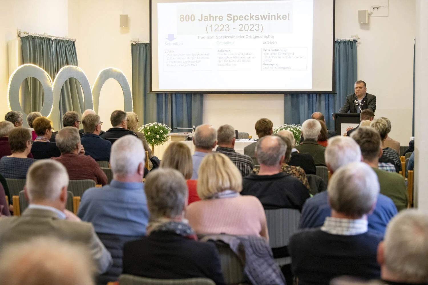 Blick über die Zuhörer zum Redner und zur Leinwand mit dem Jubiläumslogo