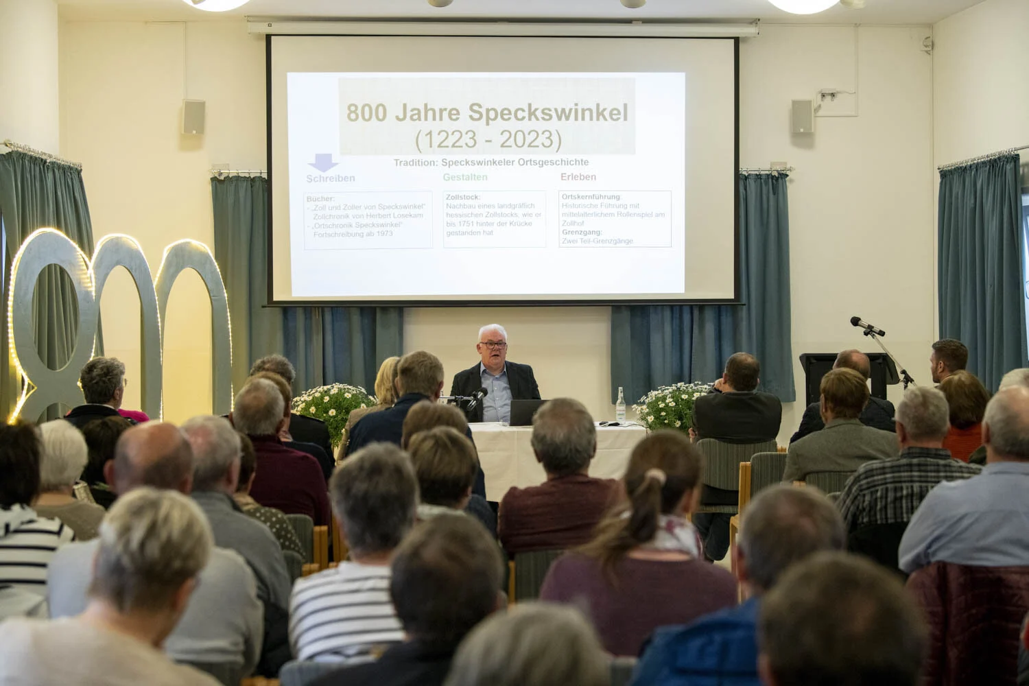 Autor sitzt am Tisch vor der Leinwand, davor Zuhörer