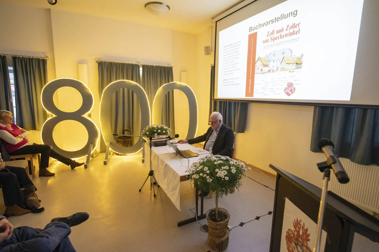 Buchvorstellung: leuchtende Zahl 800 neben der Präsentation und dem Tisch des Autors