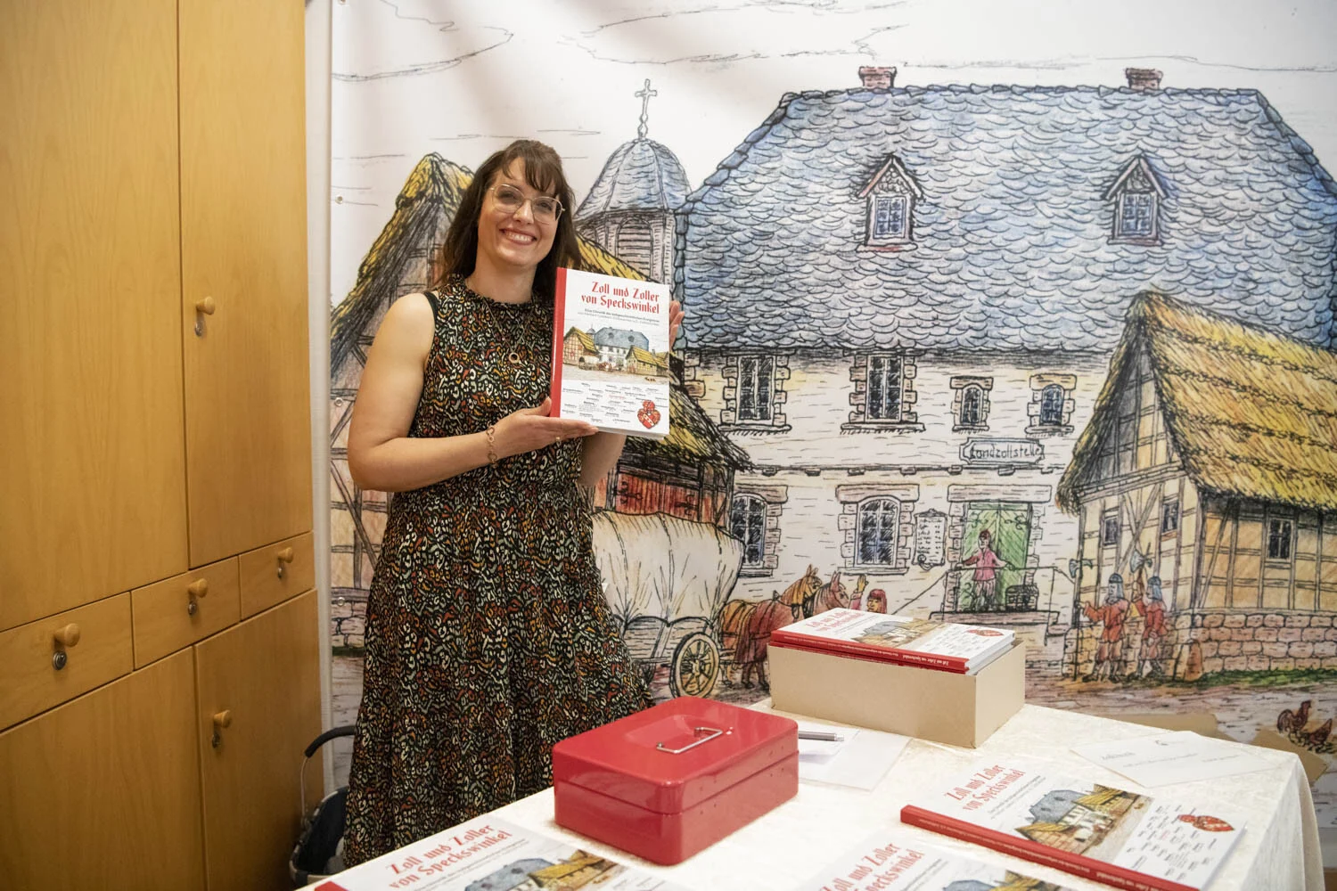 Frau zeigt das Buch vor einem großen gezeichneten Wandbild eines alten Hauses