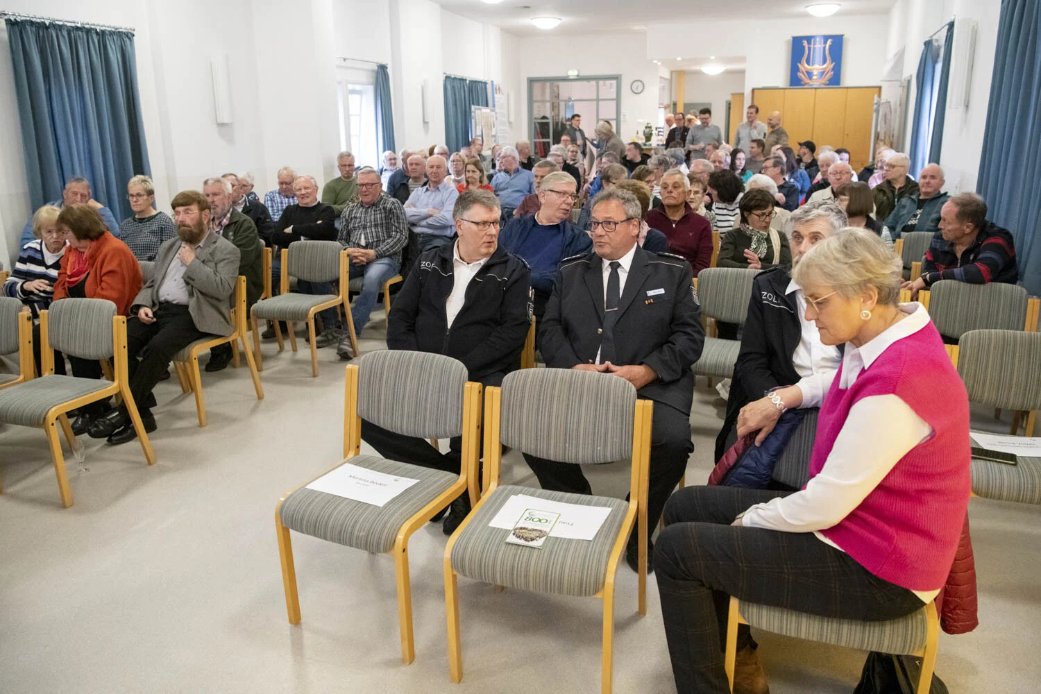 Gäste sitzen auf Stühlen im Saal, vorn eine Frau in rosa Weste