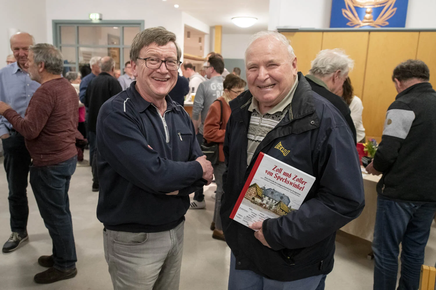 Zwei Männer lachen in die Kamera, einer hält das Buch