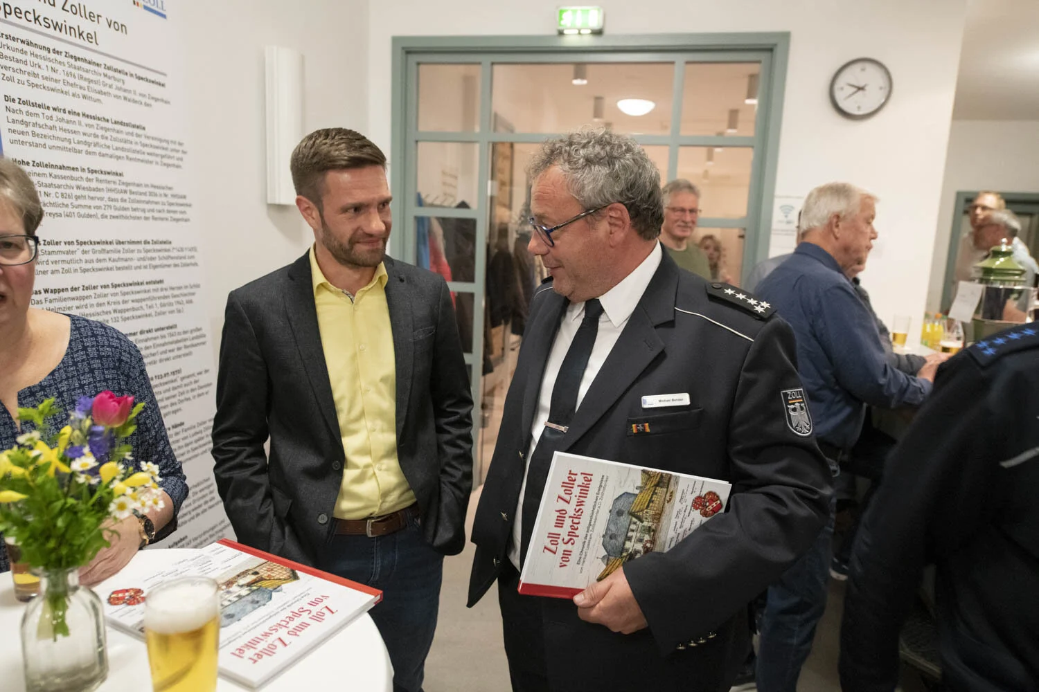 Gäste unterhalten sich am Stehtisch, ein Zollbeamter hält das Buch