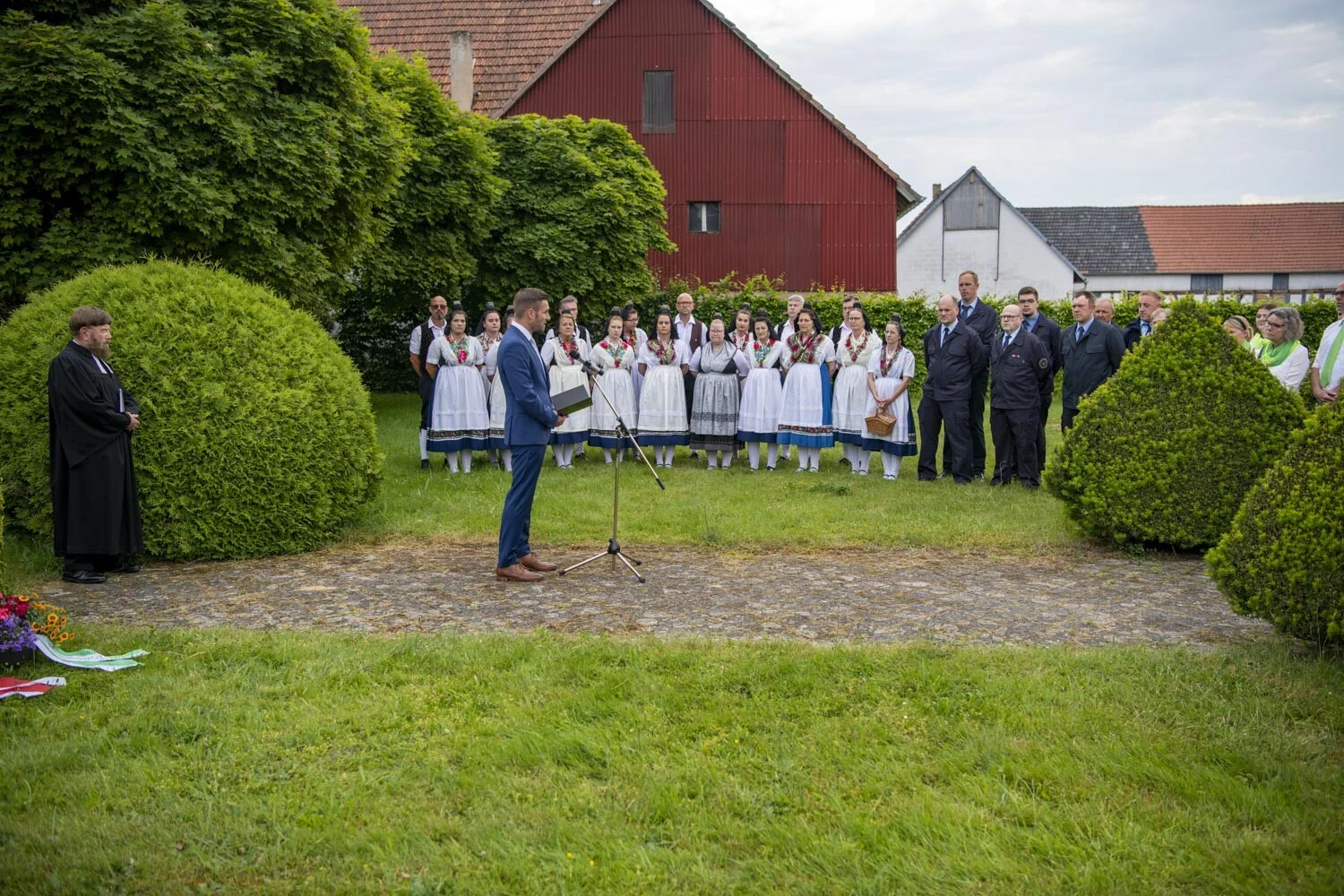 Redner und Chor in Tracht stehen auf einer Wiese zwischen runden Hecken
