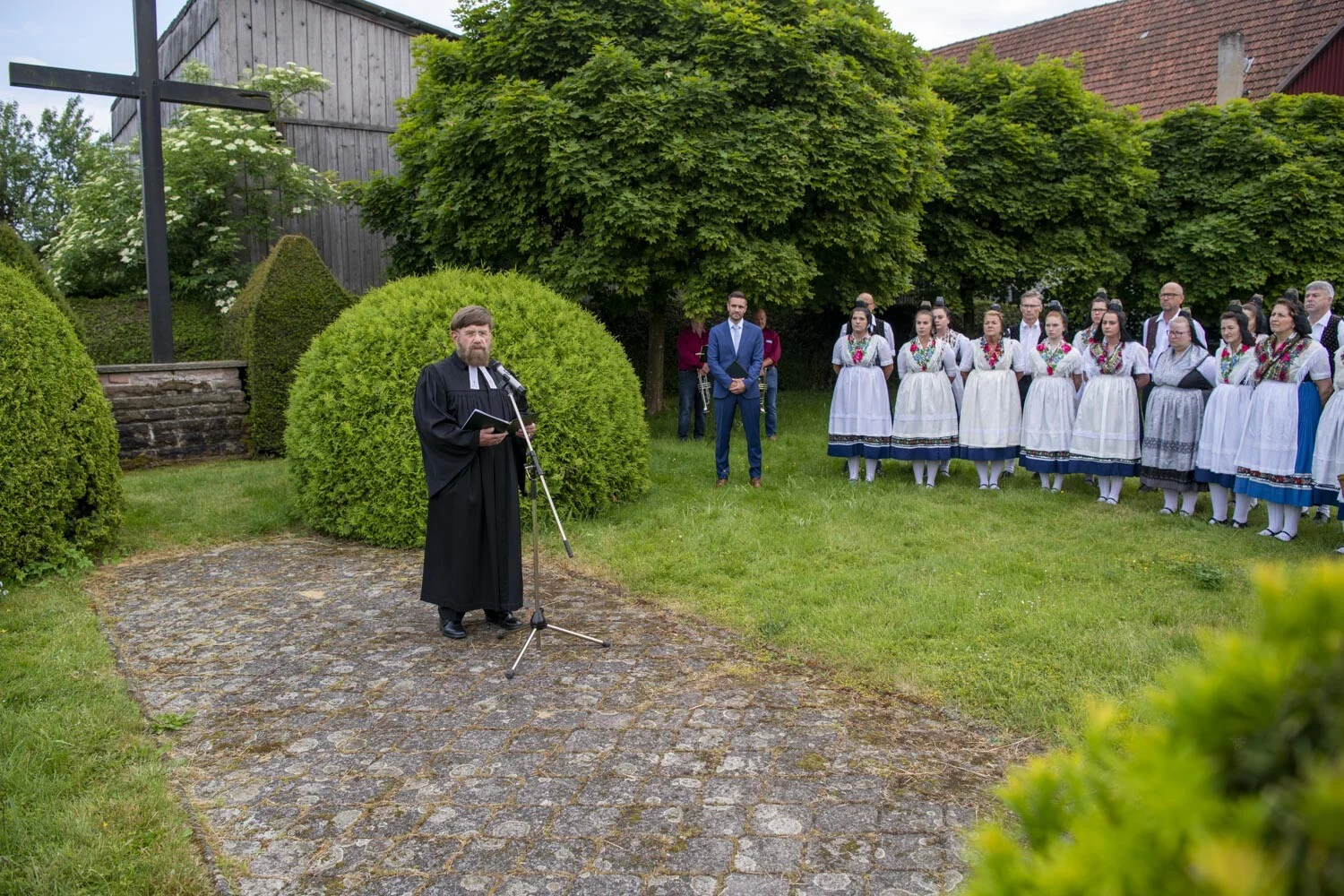 Festkommers in Speckswinkel: Geistlicher spricht am Mikrofon, dahinter ein Chor in Tracht