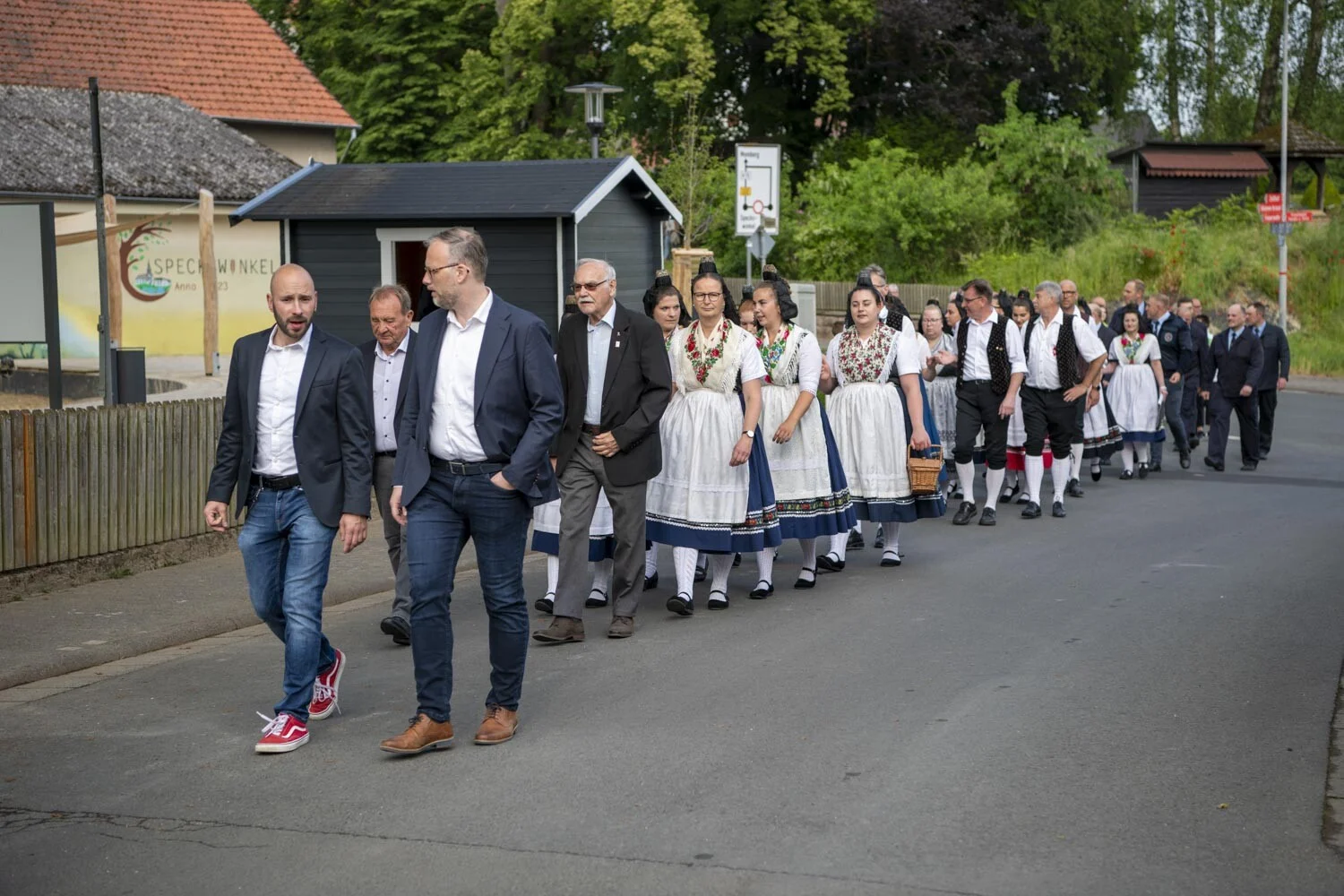 Festkommers: Ehrengäste und Trachtengruppen gehen gemeinsam eine Dorfstraße entlang