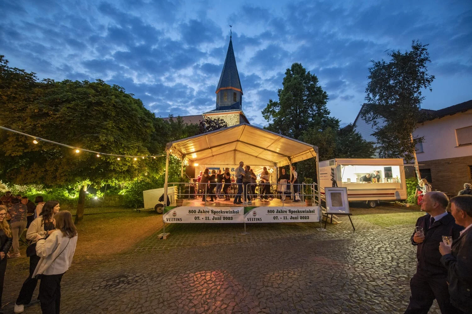 Beleuchtete Bühne unter einem Zelt, dahinter ein Kirchturm in der Abenddämmerung