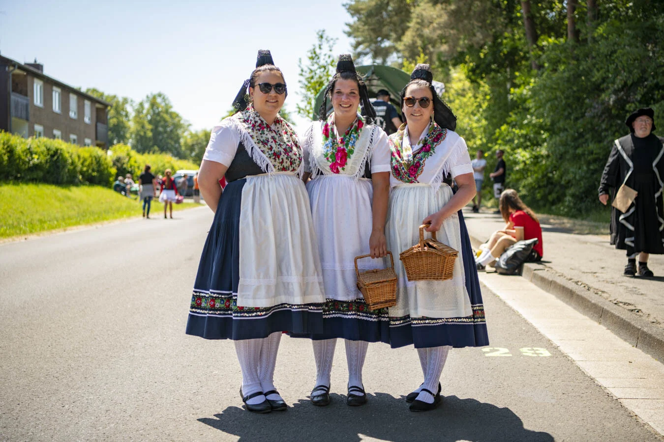 Drei Frauen in Tracht mit Körbchen stehen lächelnd am Straßenrand