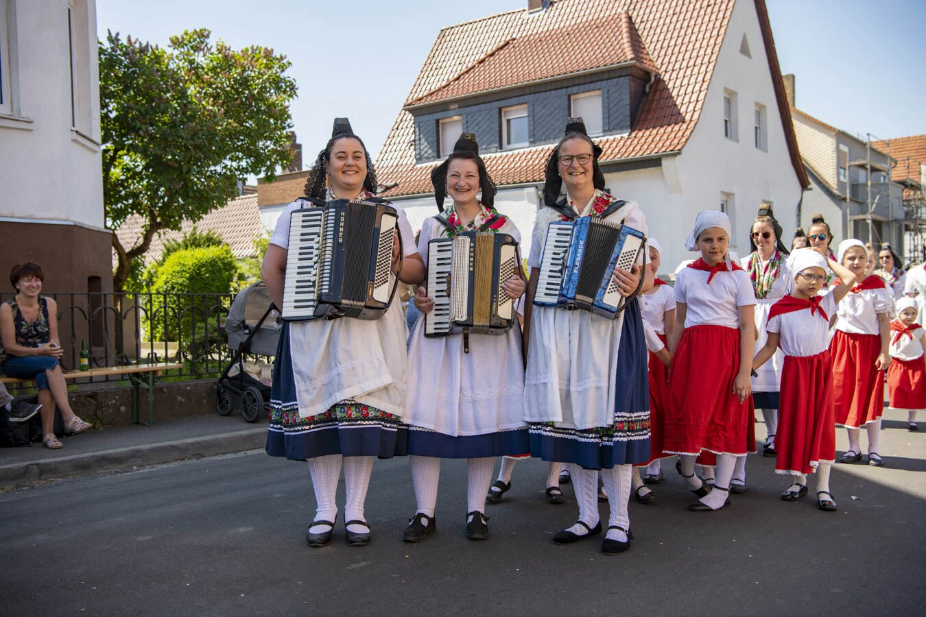 Drei Frauen in Tracht spielen Akkordeon, dahinter Kinder in roten Röcken