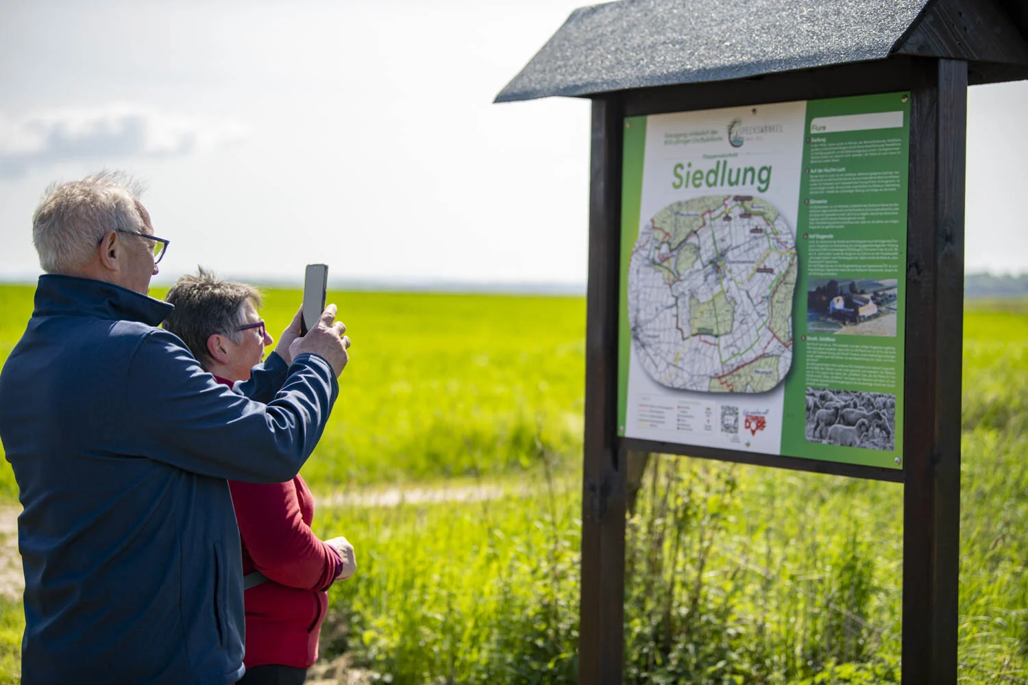 Zwei Personen fotografieren eine Infotafel mit der Überschrift Siedlung am Feldrand