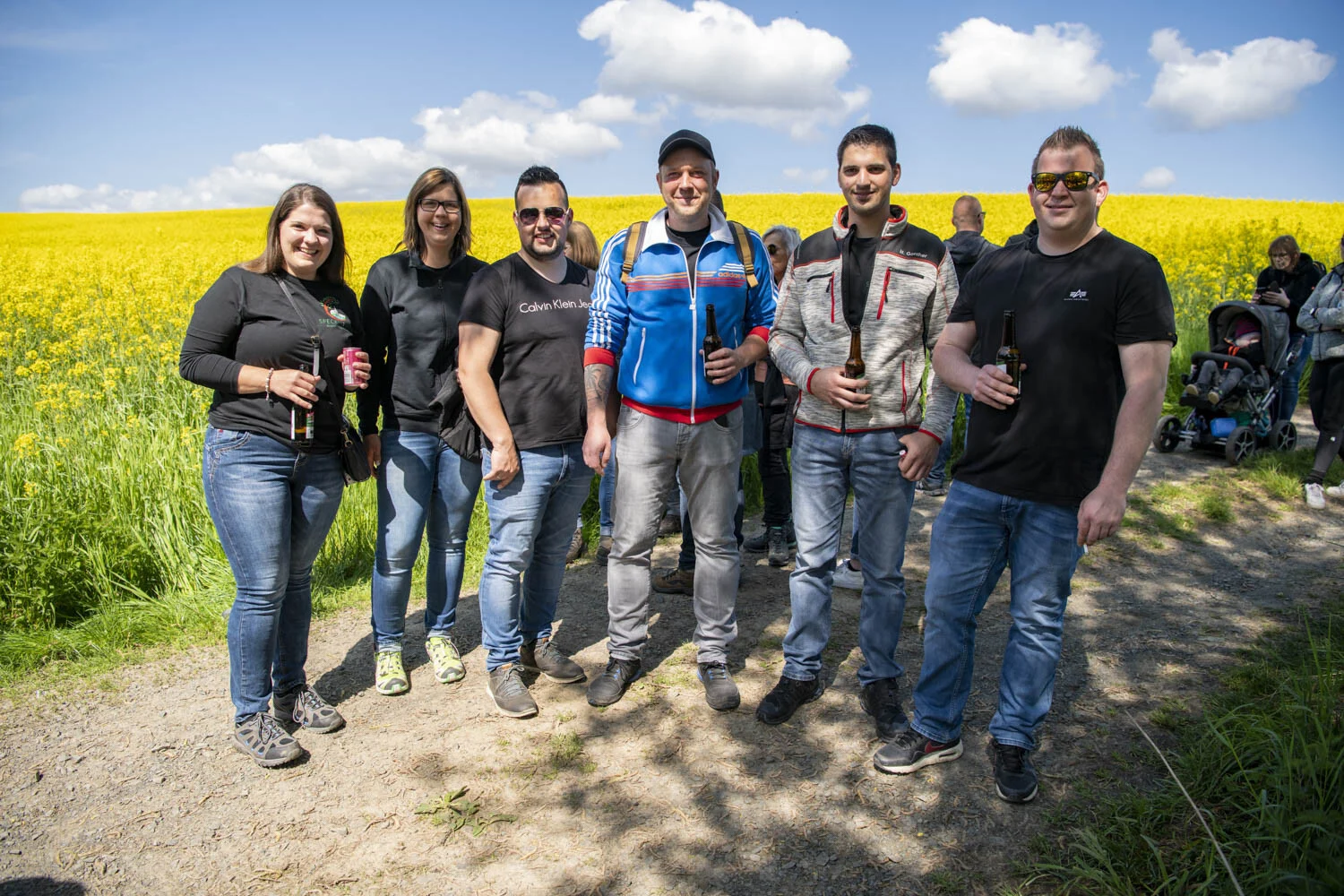 Gruppe steht mit Getränken vor einem Rapsfeld unter blauem Himmel
