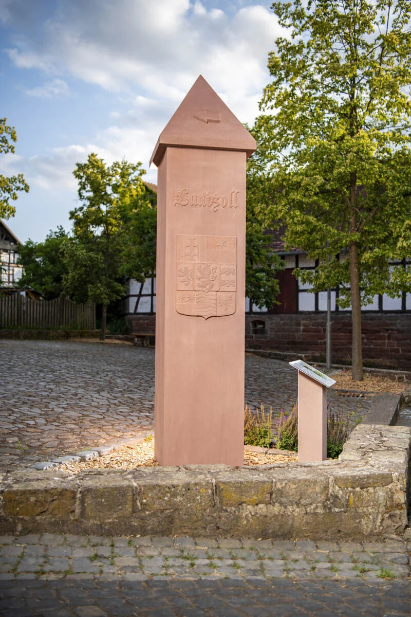 Steinpfeiler mit Pyramidenspitze vor Fachwerkhäusern, daneben eine Infotafel