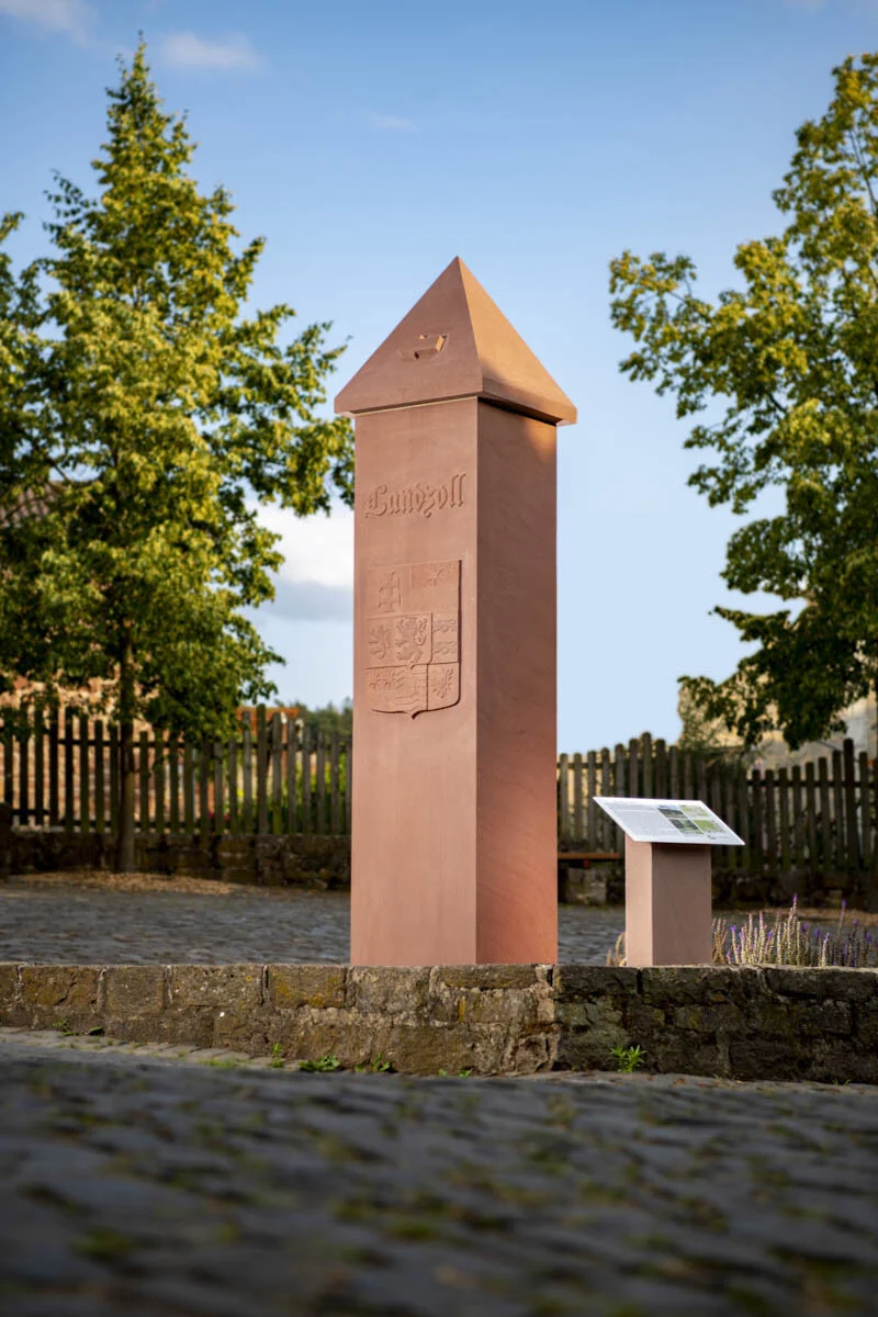 Zollstock aus rötlichem Stein vor einem Holzzaun und Bäumen im Sonnenlicht