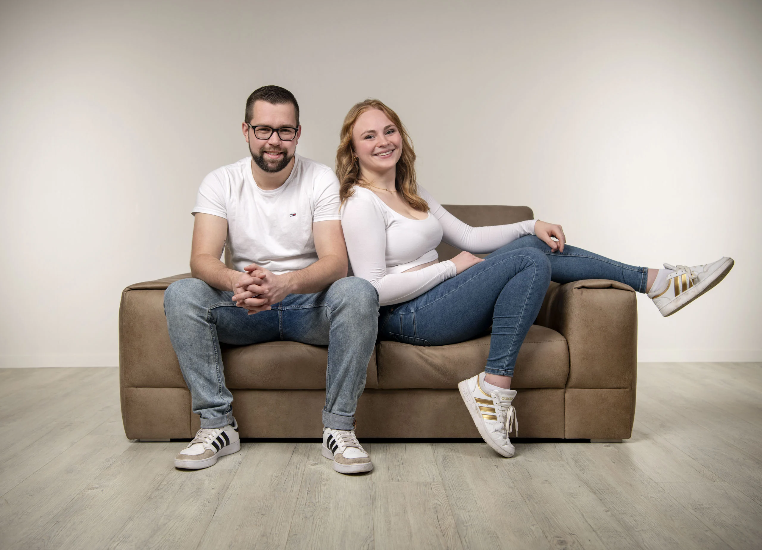 Paarfotos im Studio: Paar sitzt lächelnd auf einem braunen Sofa vor hellem Hintergrund