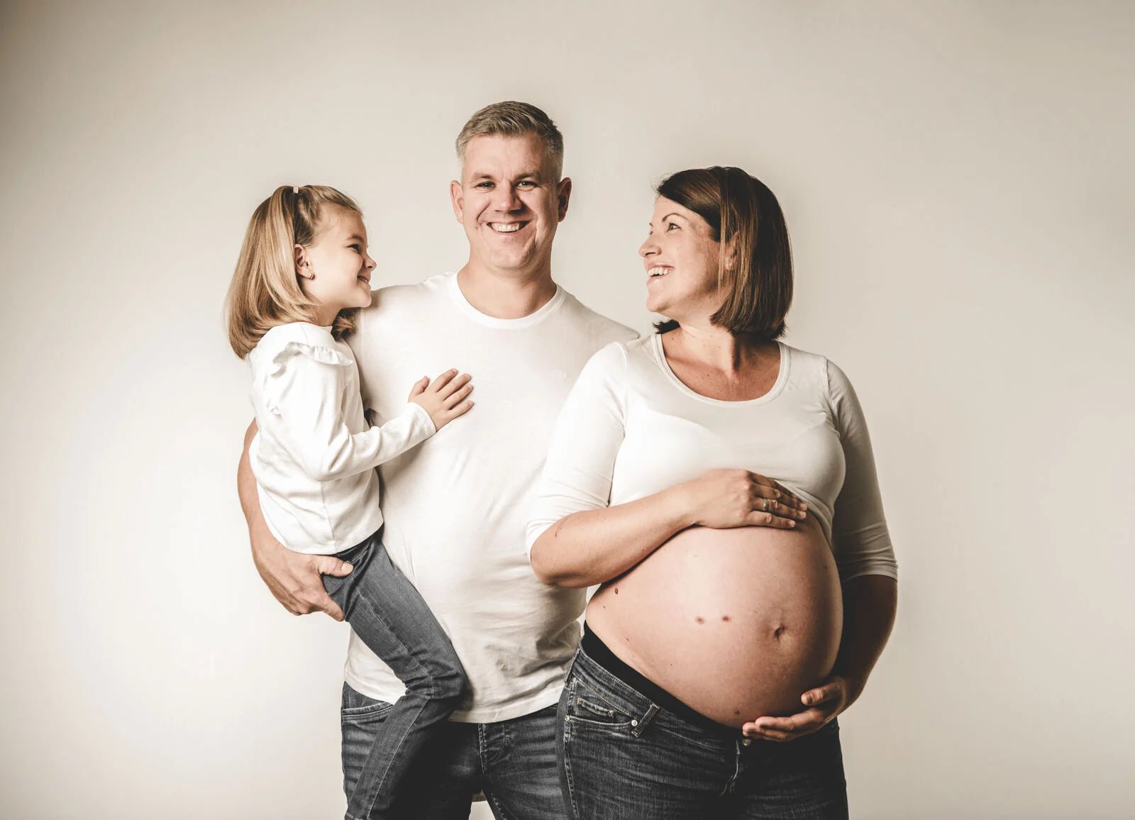 Vater hält die Tochter auf dem Arm, die Mutter hält lächelnd ihren Babybauch