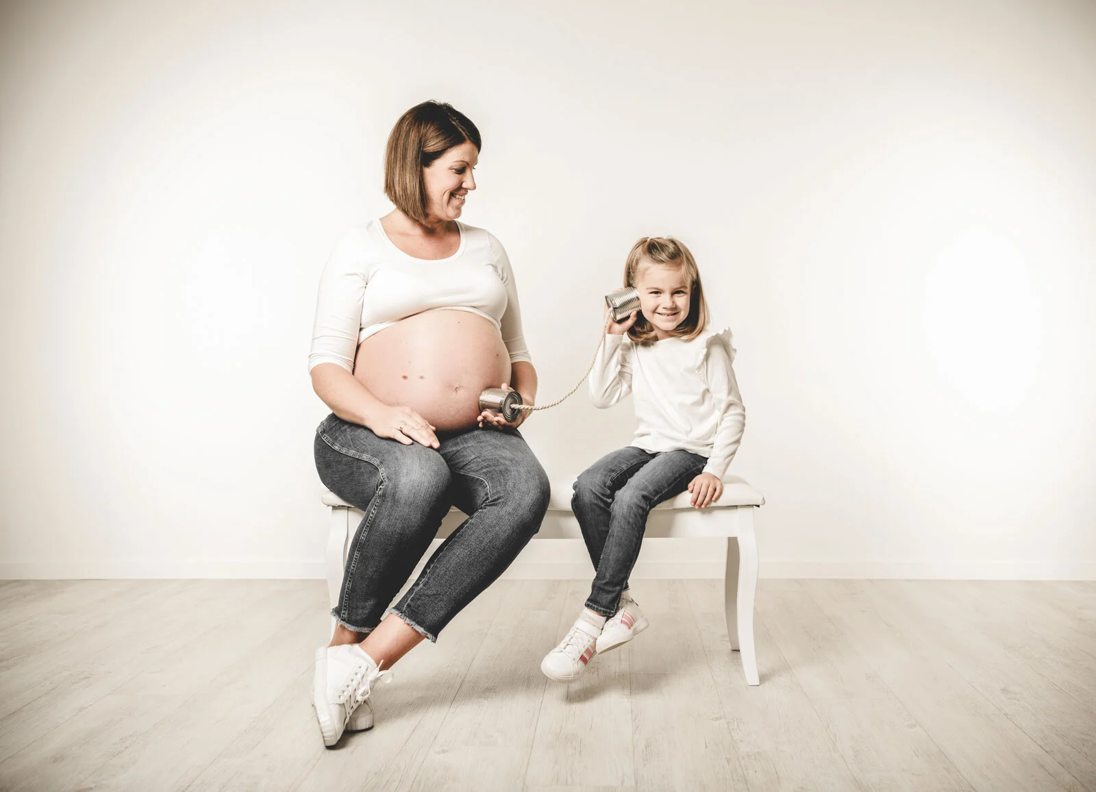 Mädchen spricht über ein Dosentelefon mit dem Babybauch der Mutter auf einer weißen Bank