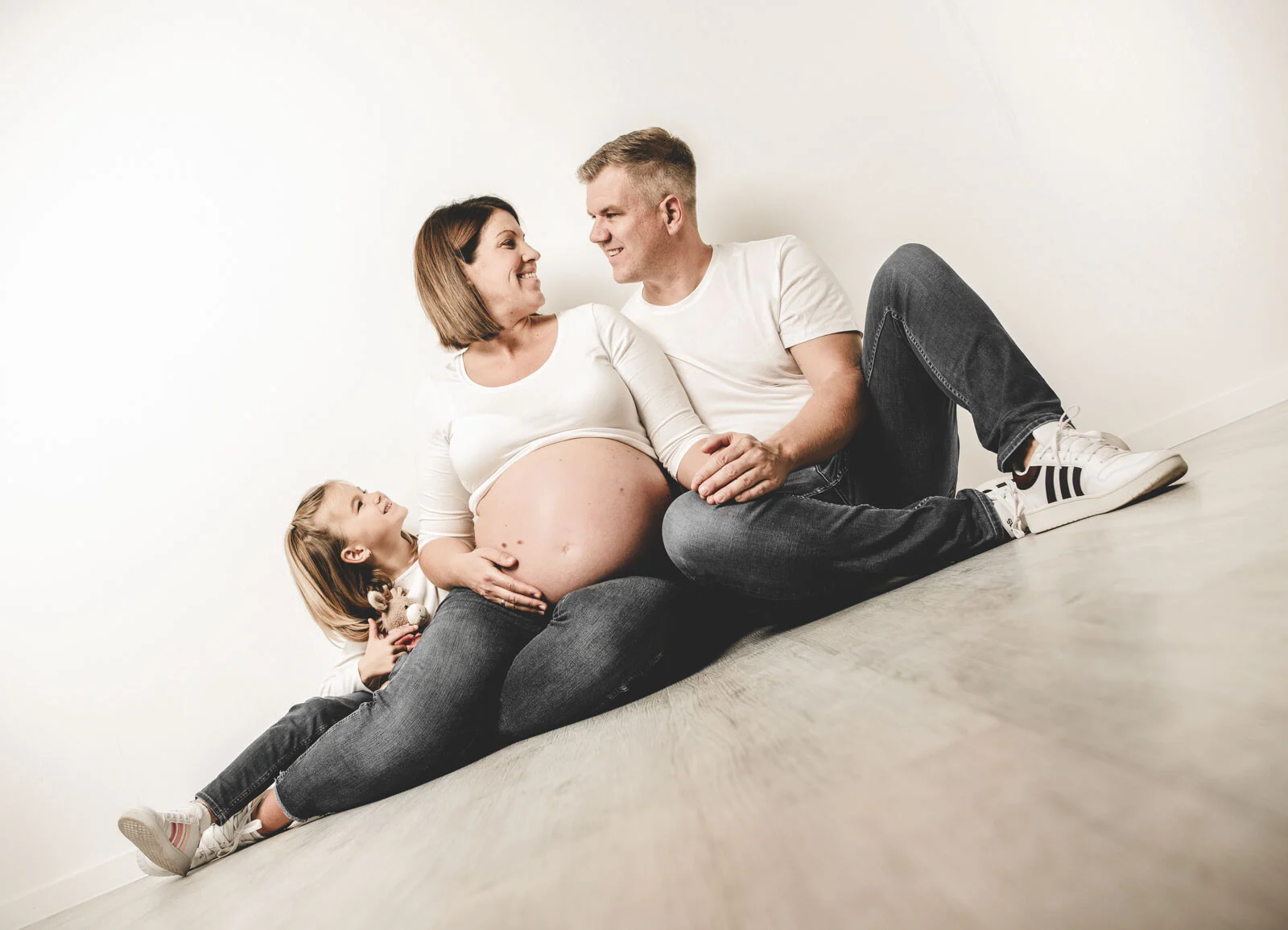 Werdende Eltern und Tochter sitzen lachend auf dem Boden vor hellem Hintergrund