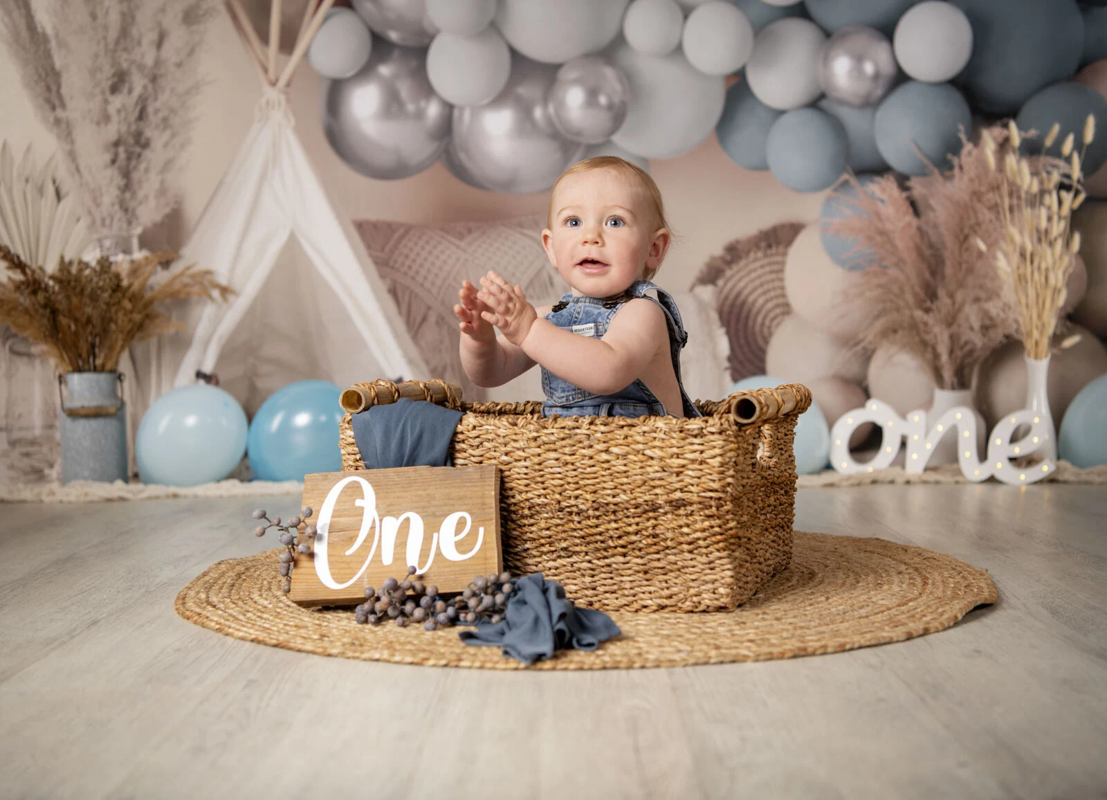 Cake Smash Fotoshooting: Kleinkind in Latzhose sitzt in einem Korb, davor ein Holzschild mit One
