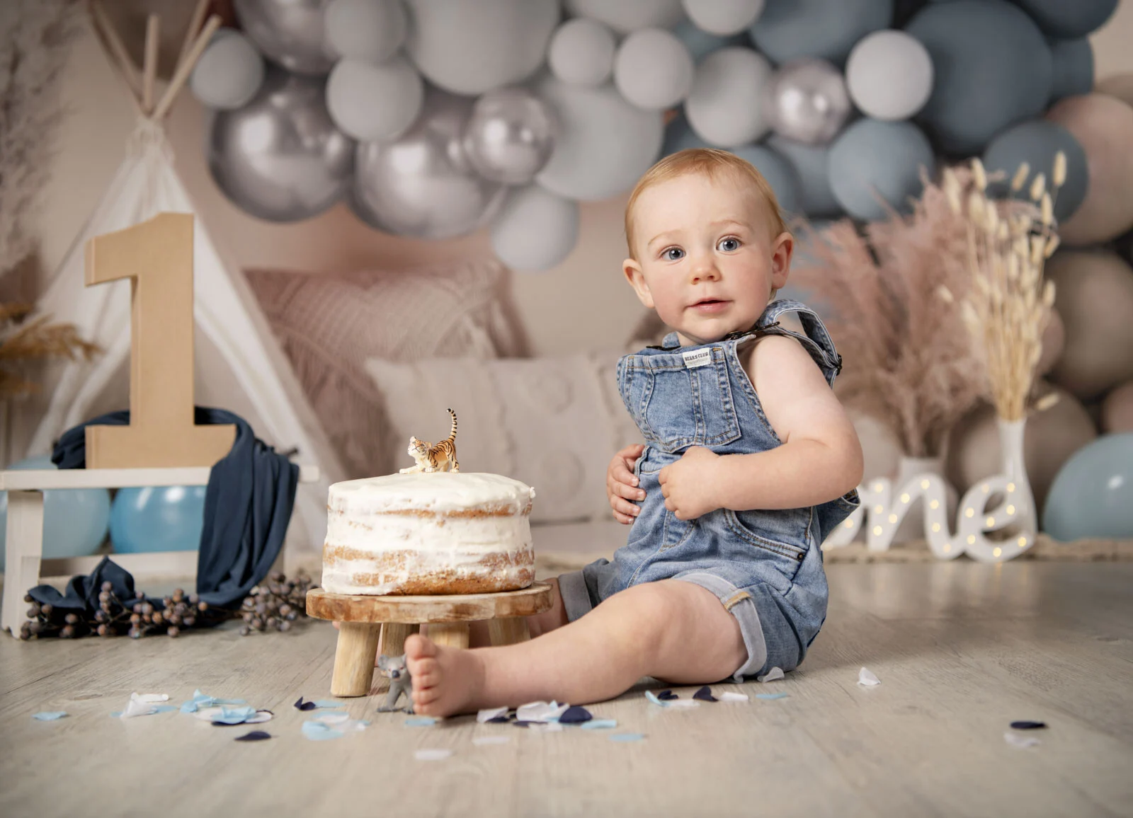 Kleinkind in Jeanslatzhose sitzt neben einer weißen Torte vor Luftballons in Blau und Silber
