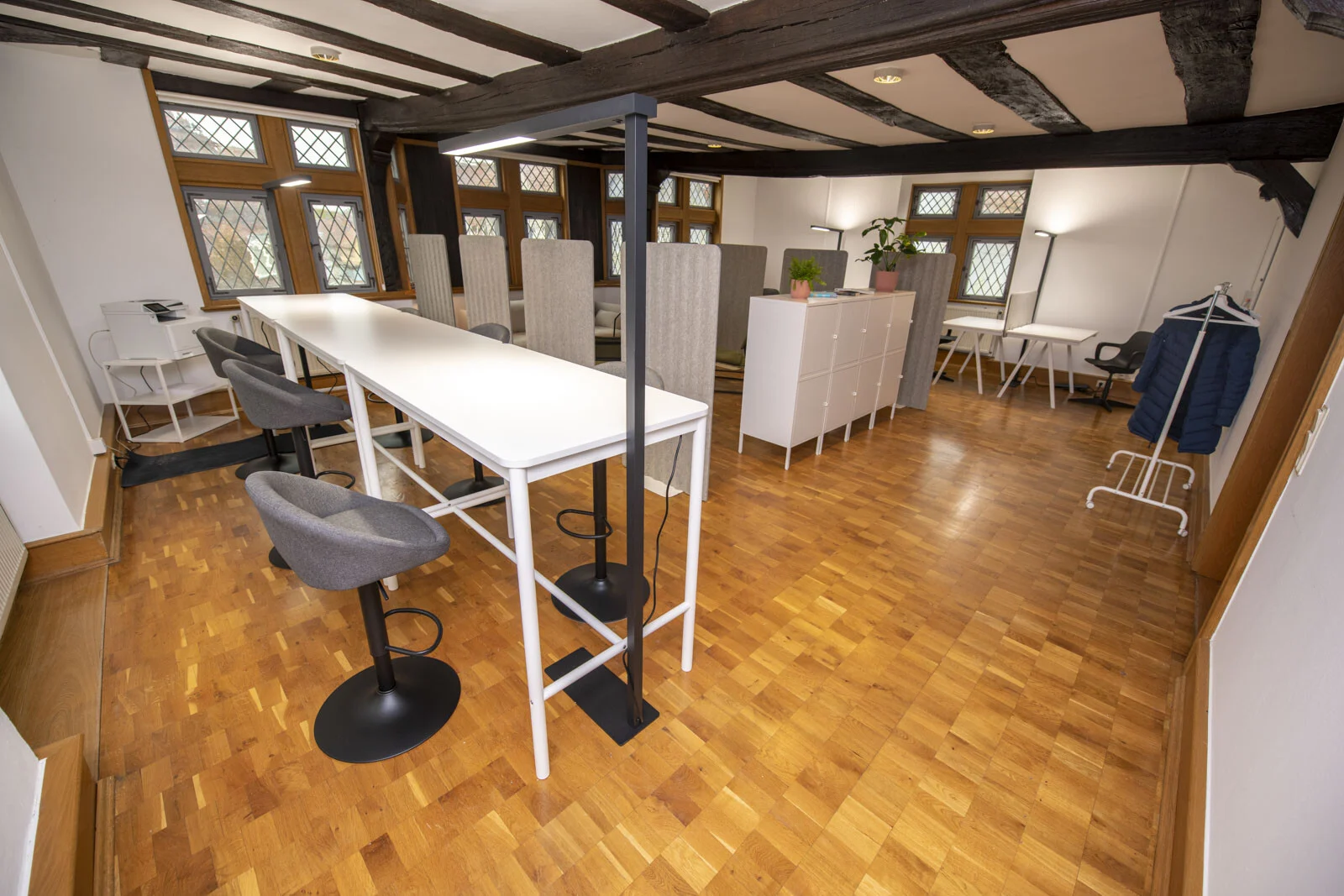 Coworking Space: lange weiße Arbeitstische mit Barhockern unter dunklen Holzbalken
