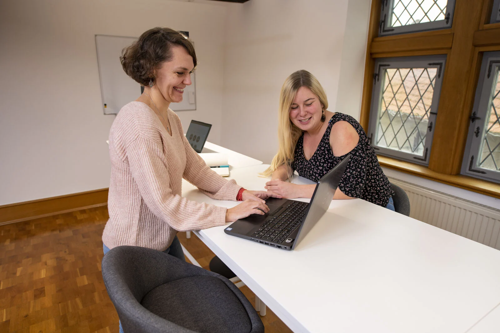 Zwei Frauen schauen gemeinsam auf einen Laptop an einem weißen Tisch