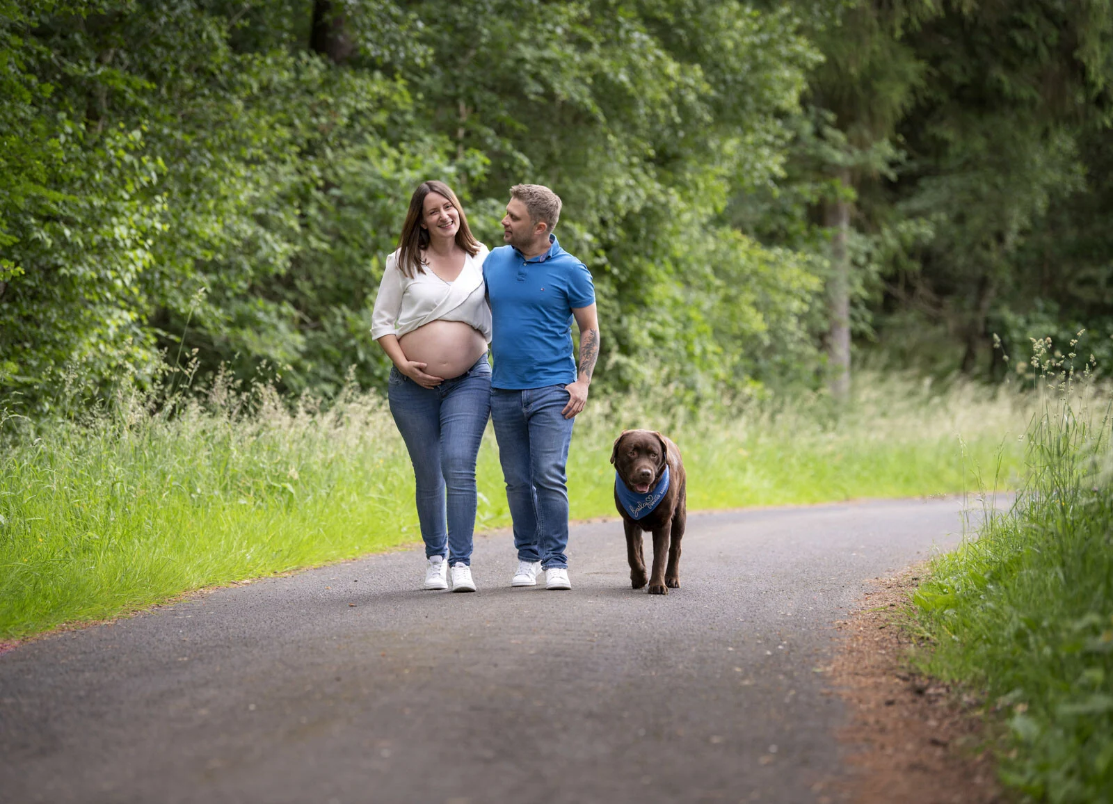 Outdoor-Fotoshooting: werdende Eltern gehen mit ihrem braunen Labrador einen Waldweg entlang