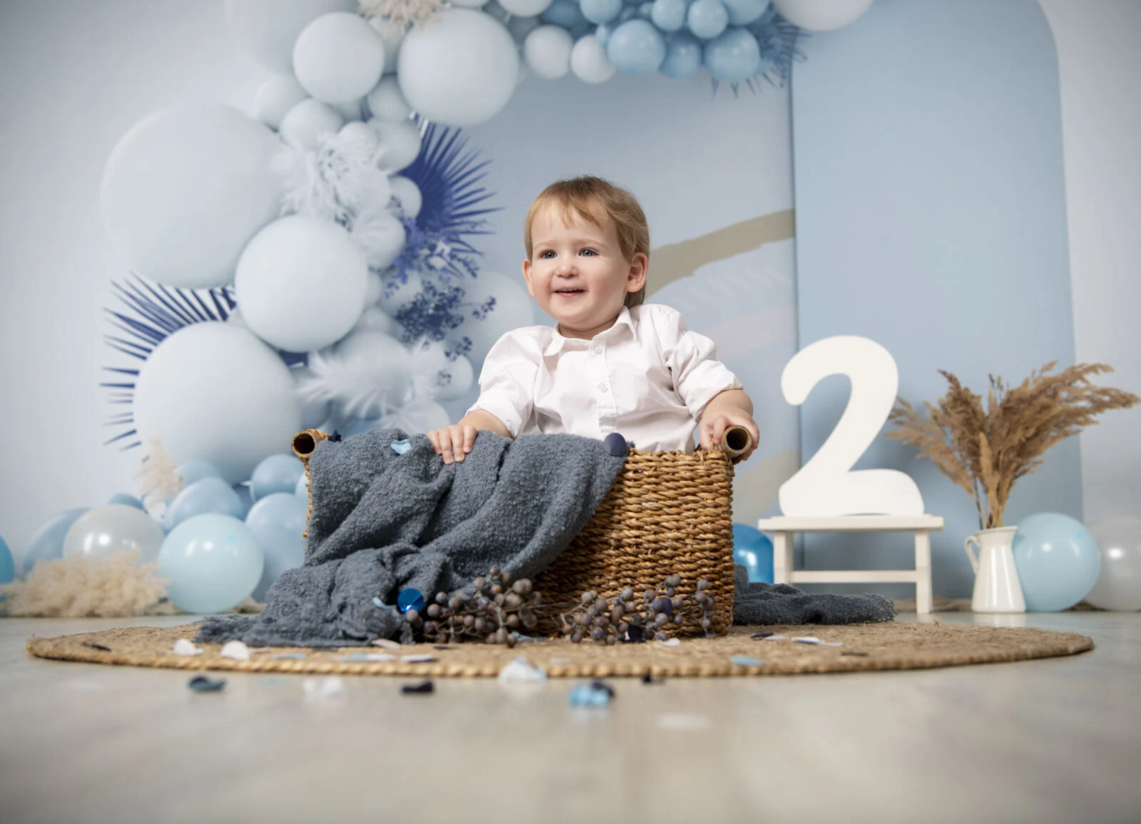 Cake Smash zum 2. Geburtstag: lachender Junge sitzt im Korb vor hellblauen Luftballons