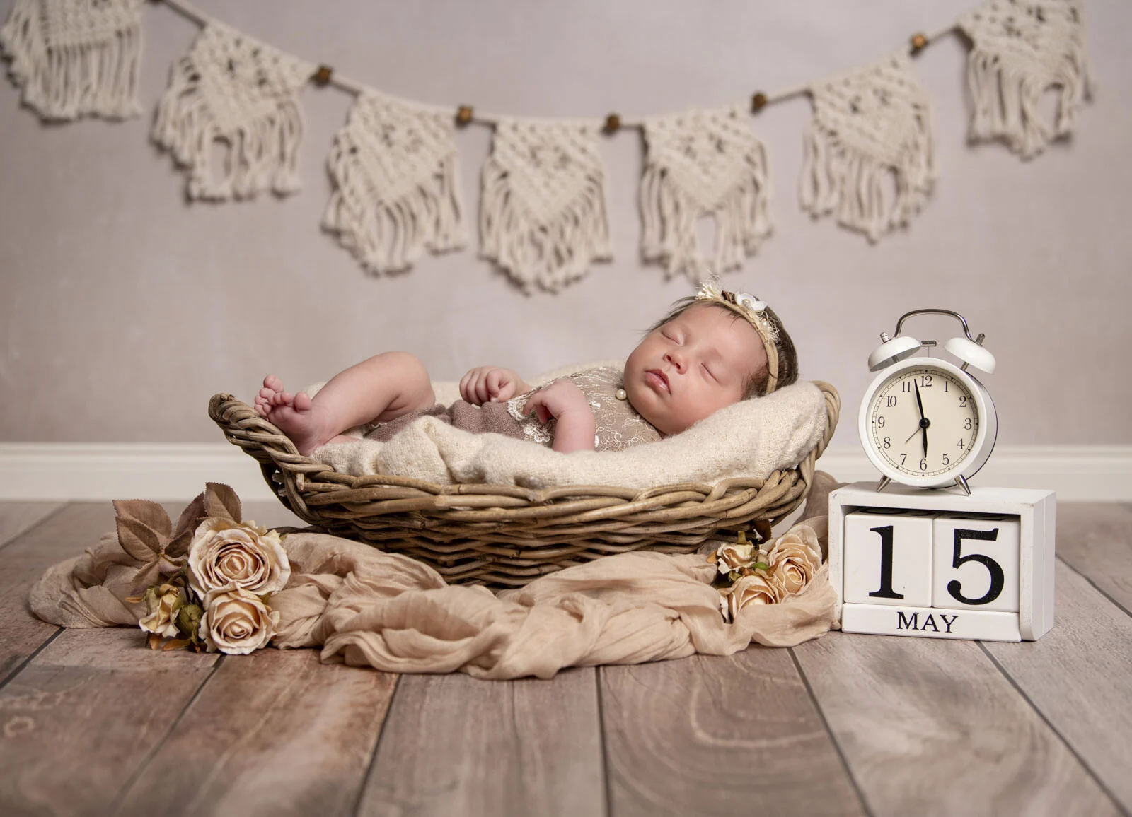 Babyfotografie: Baby schläft im Weidenkorb unter einer Makramee-Girlande, daneben ein Wecker