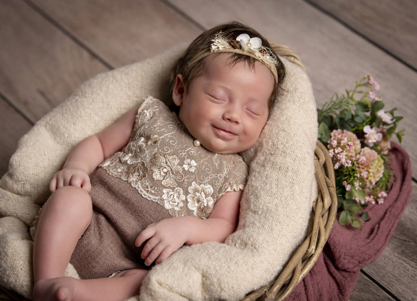 Schlafendes Baby mit Blumenhaarband lächelt im Weidenkorb, daneben kleine Blumen