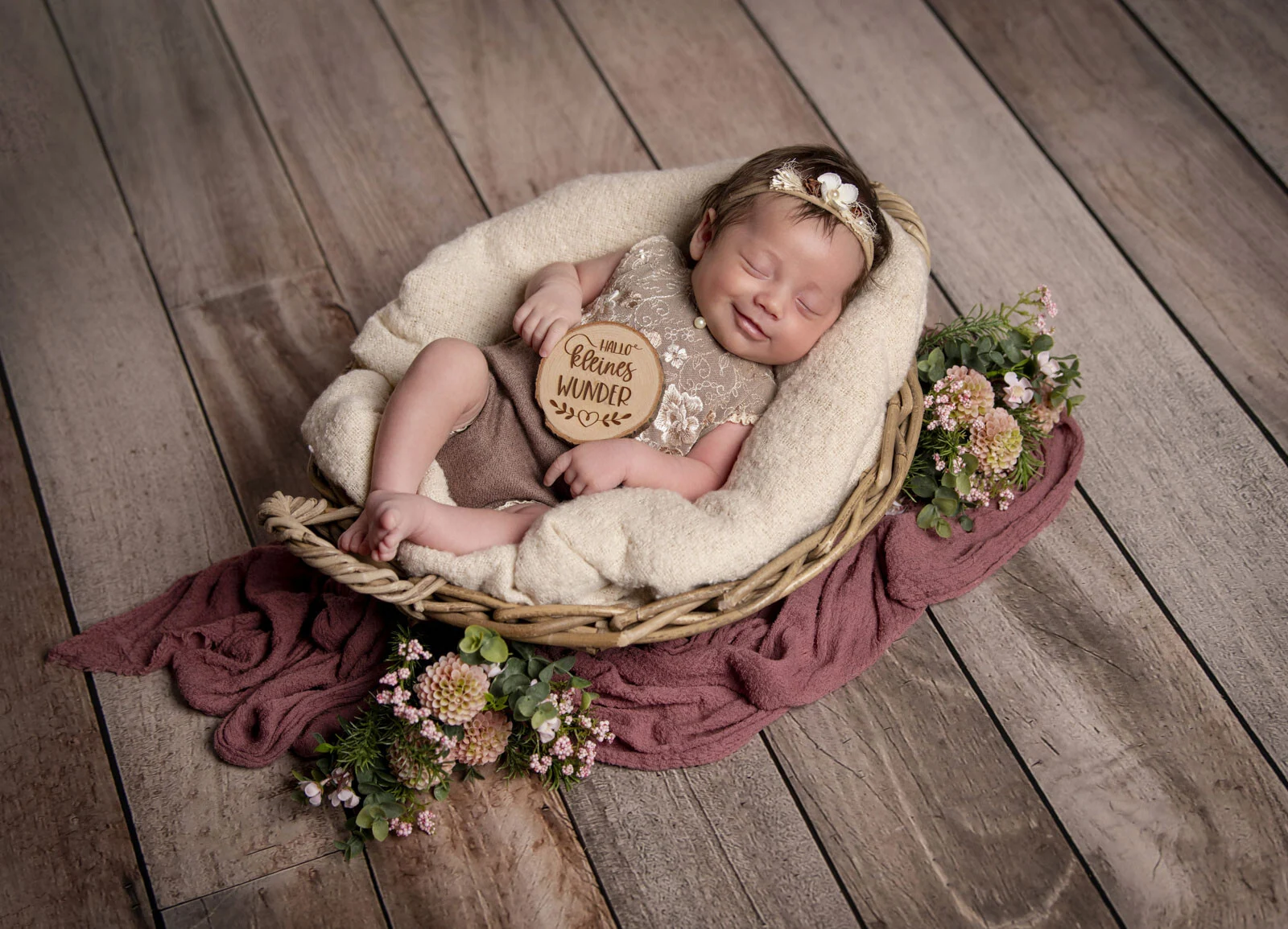 Baby liegt mit einem runden Holzschild im Korb auf einem rosa Tuch, umgeben von Blumen