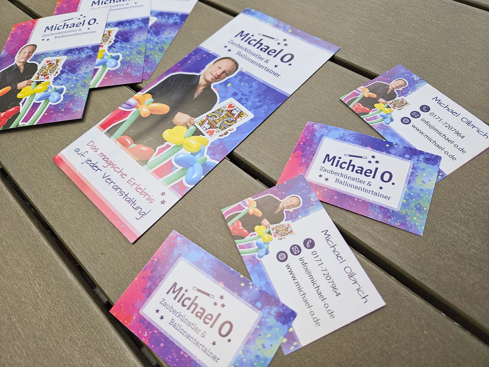Autogrammkarten, Flyer und Visitenkarten von Michael O. in Lila und Pink auf einem Holztisch