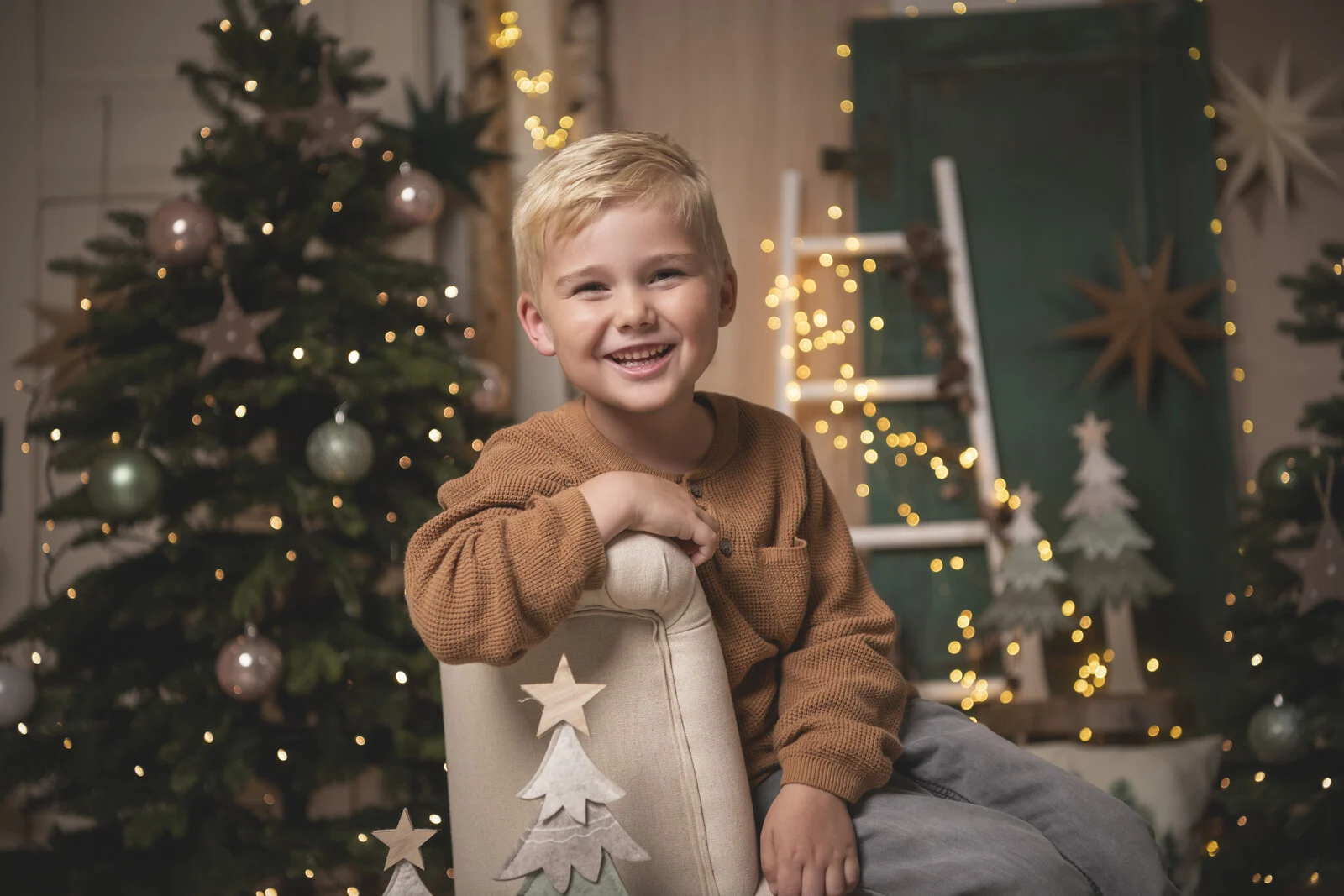 Blonder Junge im braunen Pulli lehnt lachend über einem hellen Sessel vor dem Tannenbaum