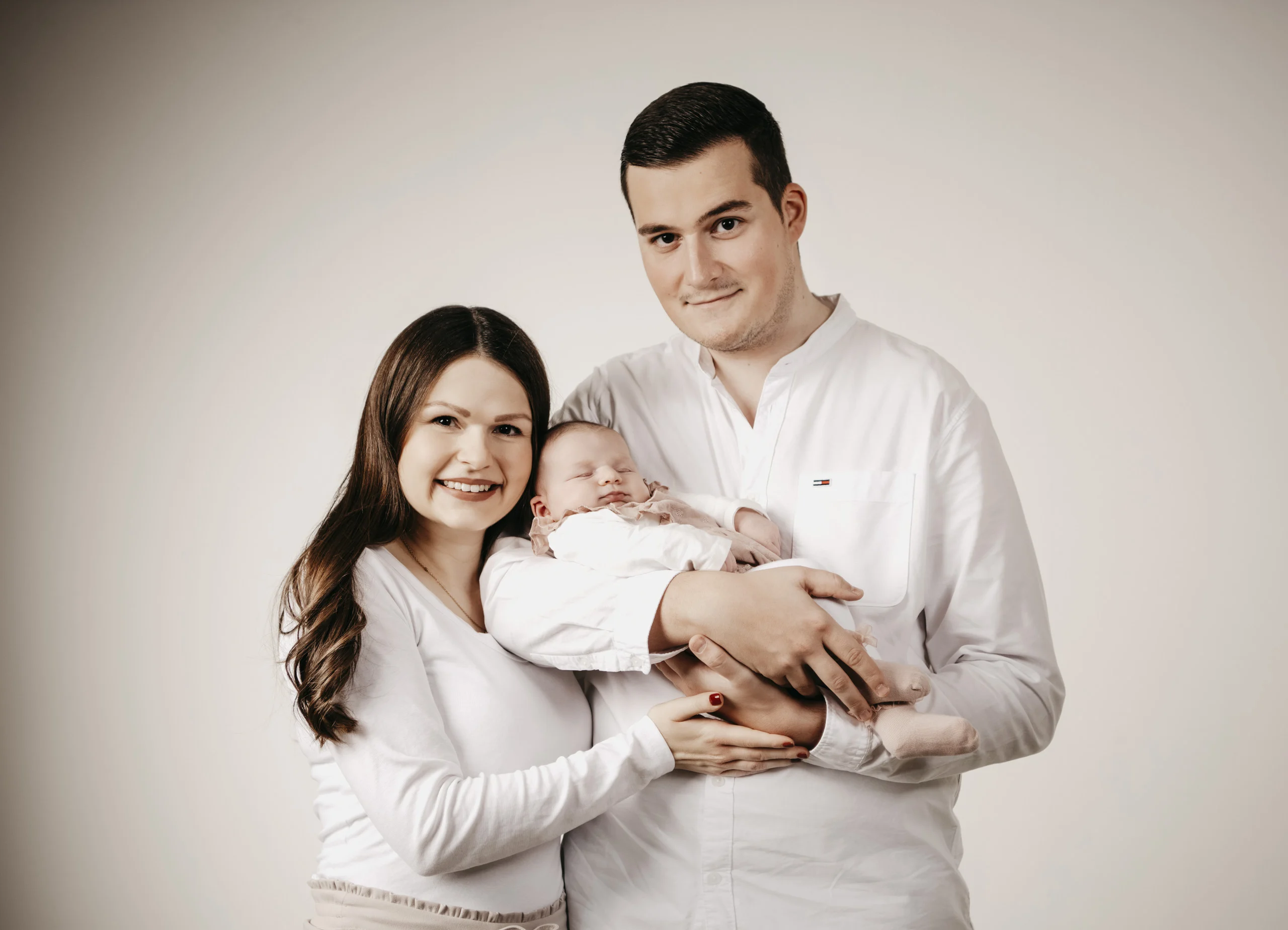 Familienfotos mit Baby: junges Paar in Weiß hält lächelnd sein Neugeborenes