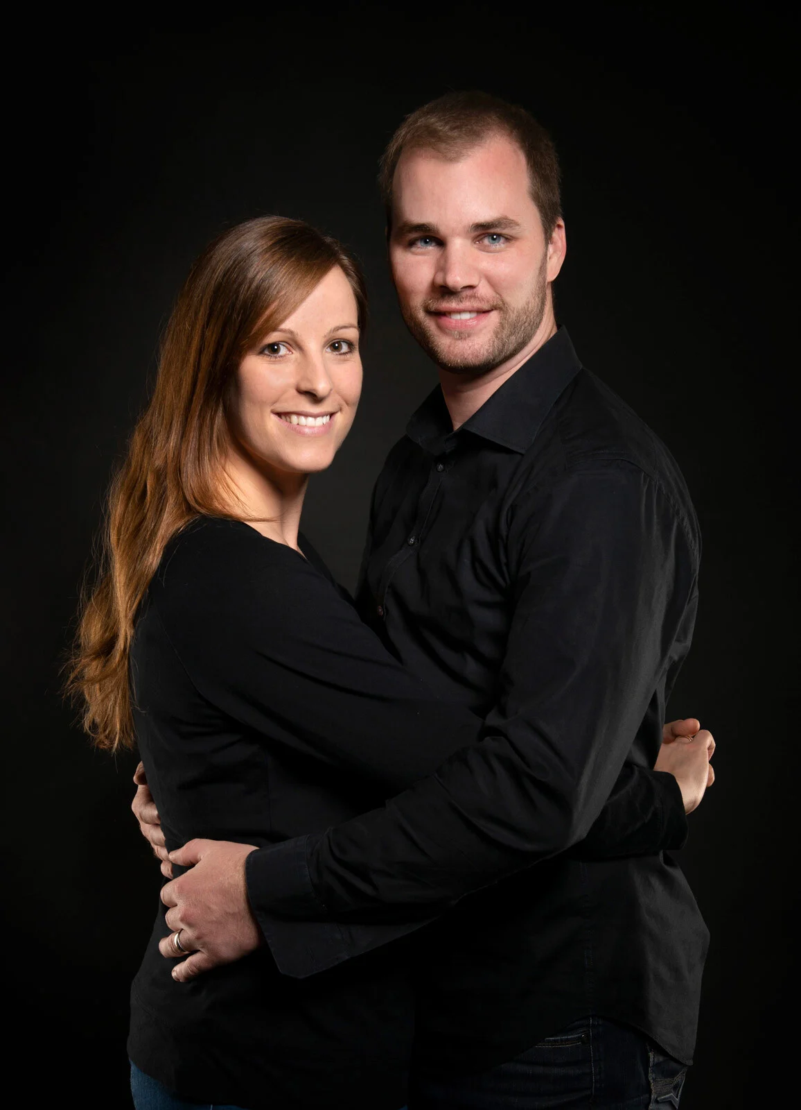 Generationen-Fotoshooting: junges Paar | Peter Lauritis Photography Junges Paar in schwarzer Kleidung steht eng umarmt vor dunklem Hintergrund