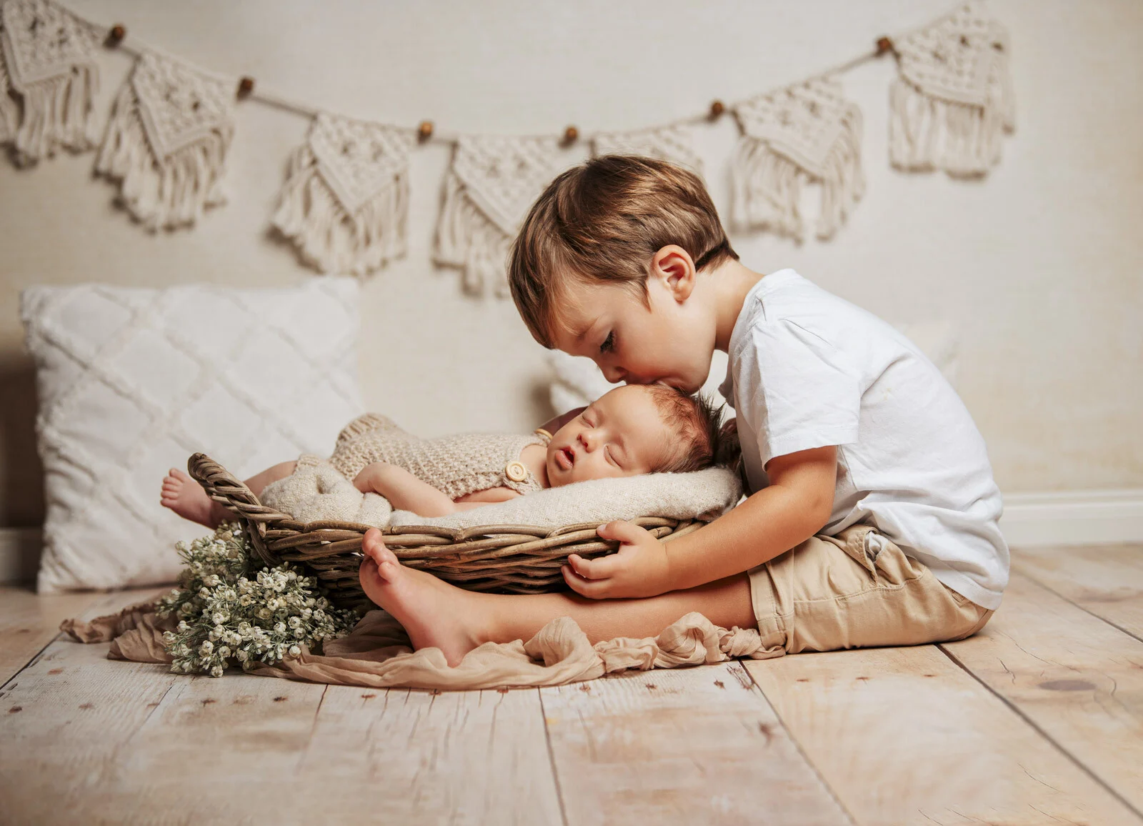 Neugeborenes im Korb mit kleinem Jungen | Peter Lauritis Photography Kleiner Junge küsst ein schlafendes Neugeborenes im Weidenkorb, dahinter eine Makramee-Girlande – Neugeborenenfotos im Fotostudio
