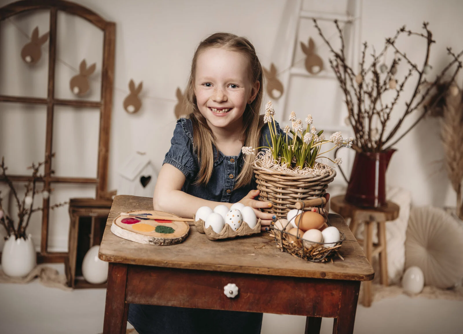 Lachendes Mädchen sitzt am Holztisch mit Ostereiern und Frühlingsblumen