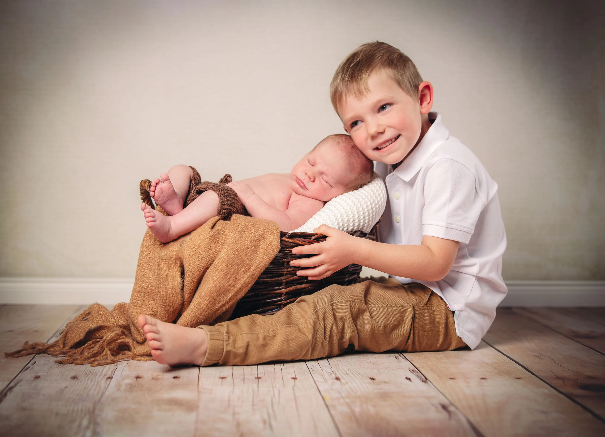 Babyfotos Neugeborene Fotogalerie 006 | Peter Lauritis Photography Junge sitzt neben einem Korb, in dem ein Neugeborenes schläft, und lächelt