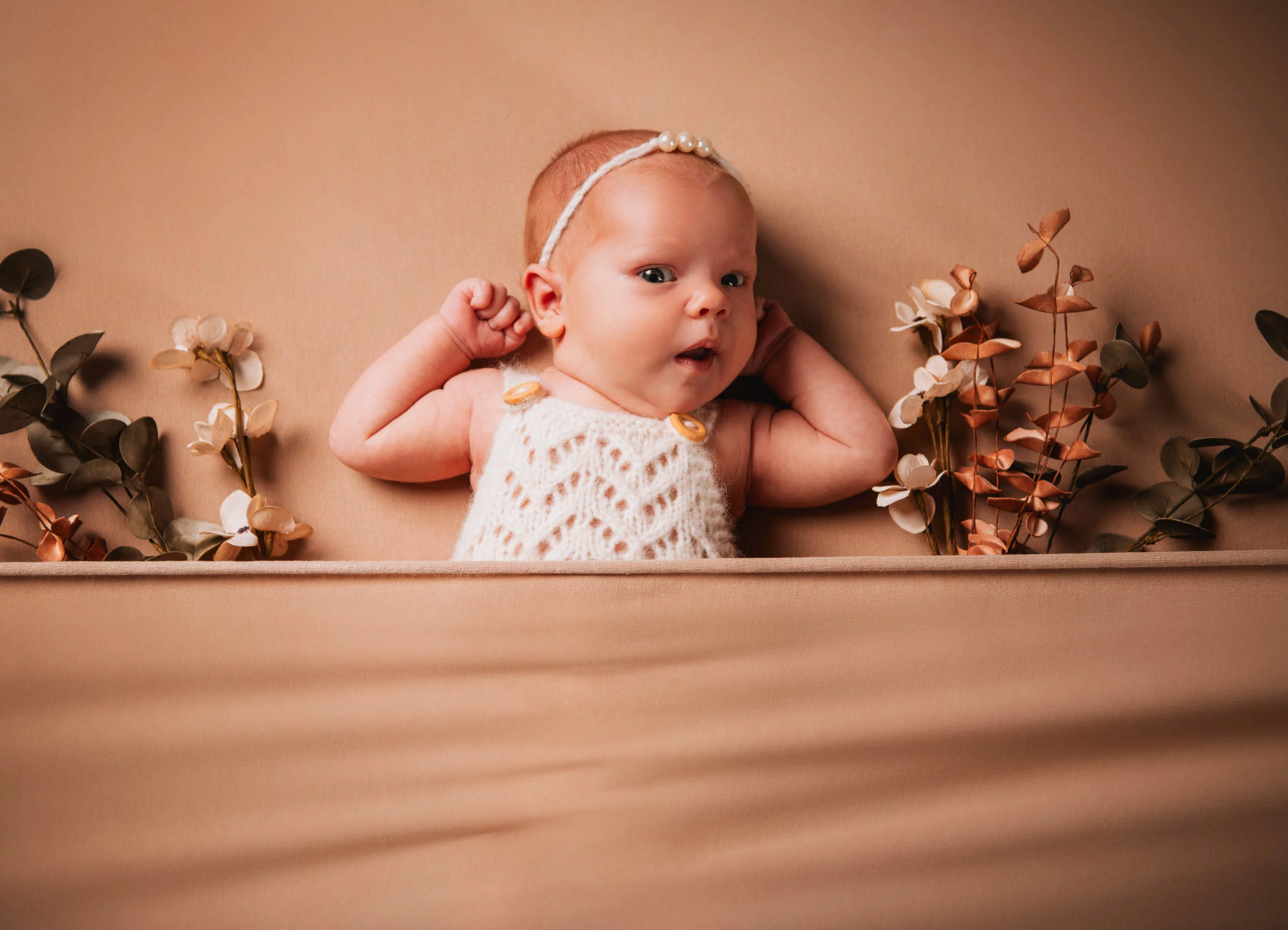 Babyfotos Neugeborene Fotogalerie 014 | Peter Lauritis Photography Baby mit Haarband liegt zwischen Trockenblumen auf beigem Stoff