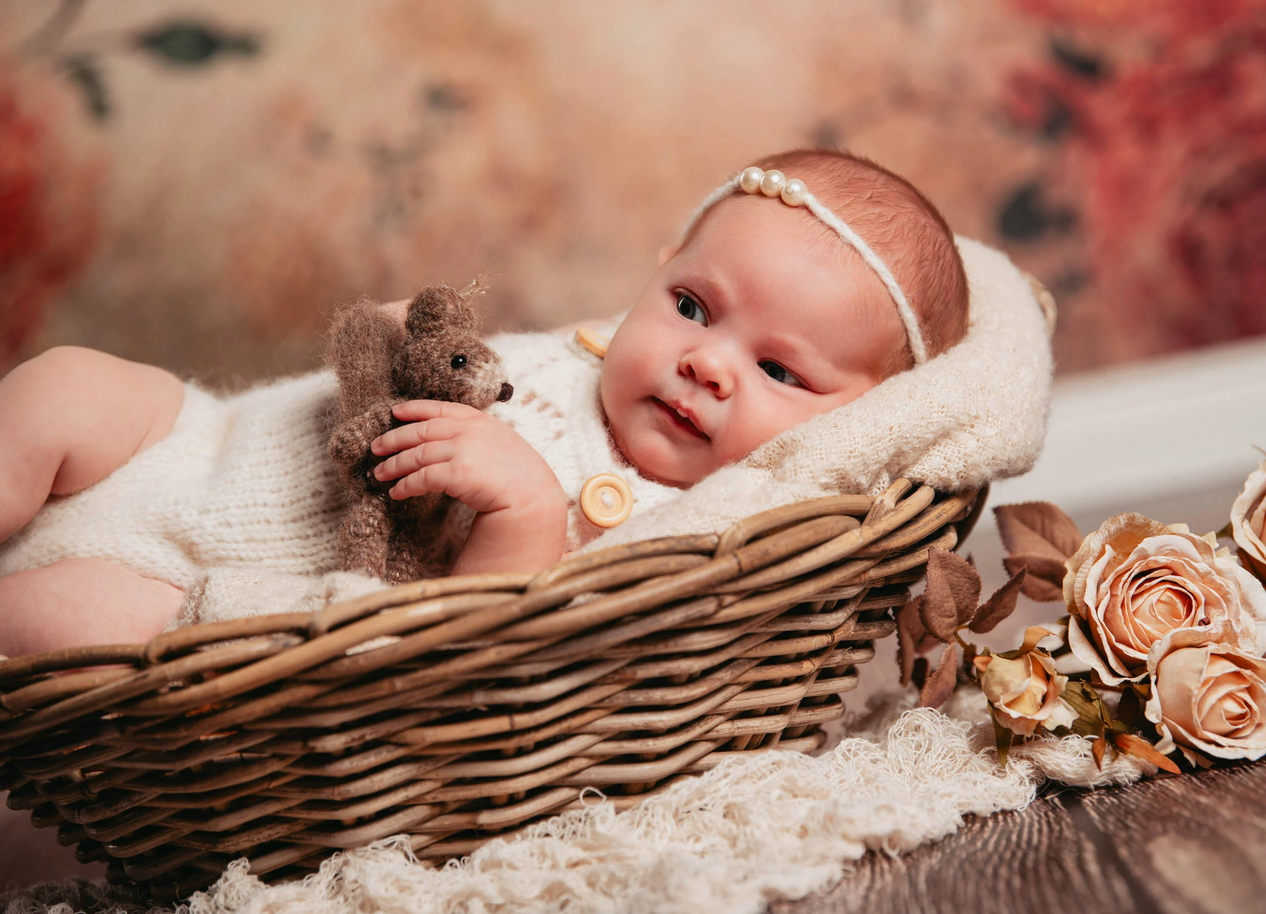 Babyfotos Neugeborene Fotogalerie 016 | Peter Lauritis Photography Baby mit Haarband hält einen kleinen Teddy im Korb vor Rosenhintergrund