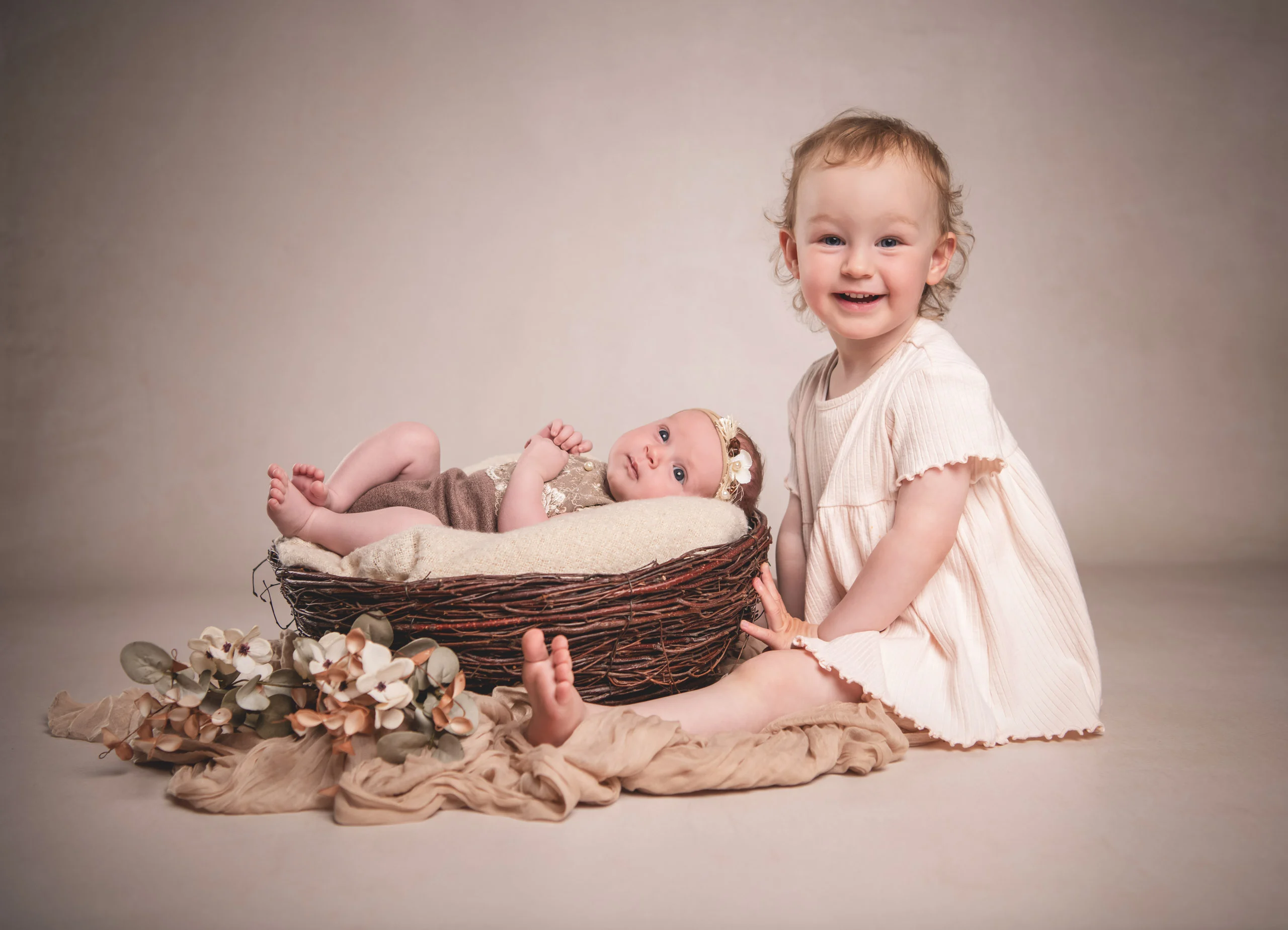 Babyfotos Neugeborene Fotogalerie 022 | Peter Lauritis Photography Kleinkind im hellen Kleid sitzt lachend neben einem Korb mit Baby