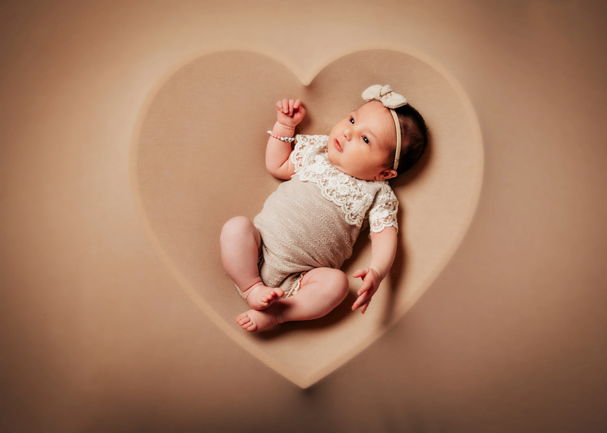 Babyfotos Neugeborene Fotogalerie 031 | Peter Lauritis Photography Baby im Spitzenbody liegt in einer herzförmigen Mulde auf beigem Hintergrund