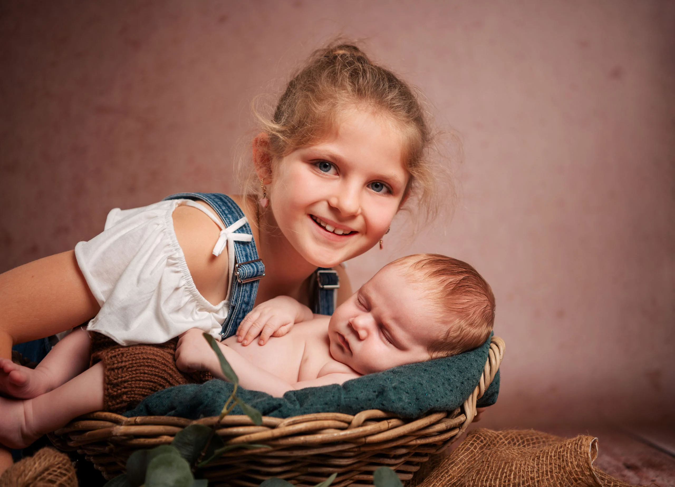 Babyfotos Neugeborene Fotogalerie 043 | Peter Lauritis Photography Mädchen in Latzhose lächelt neben dem schlafenden Baby im Korb