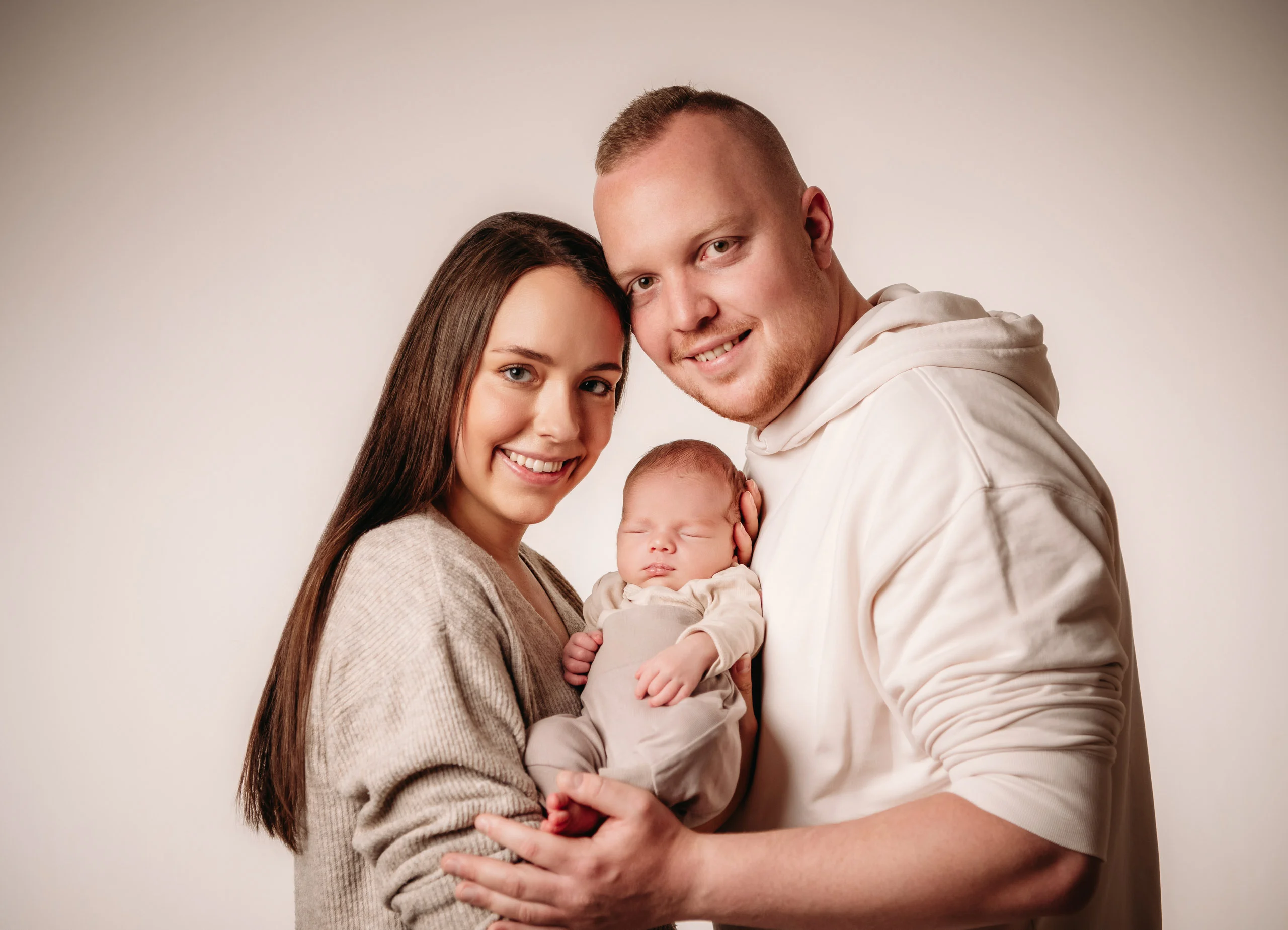 Babyfotos Neugeborene Fotogalerie 064 | Peter Lauritis Photography Lächelndes Paar hält sein Neugeborenes vor hellem Hintergrund