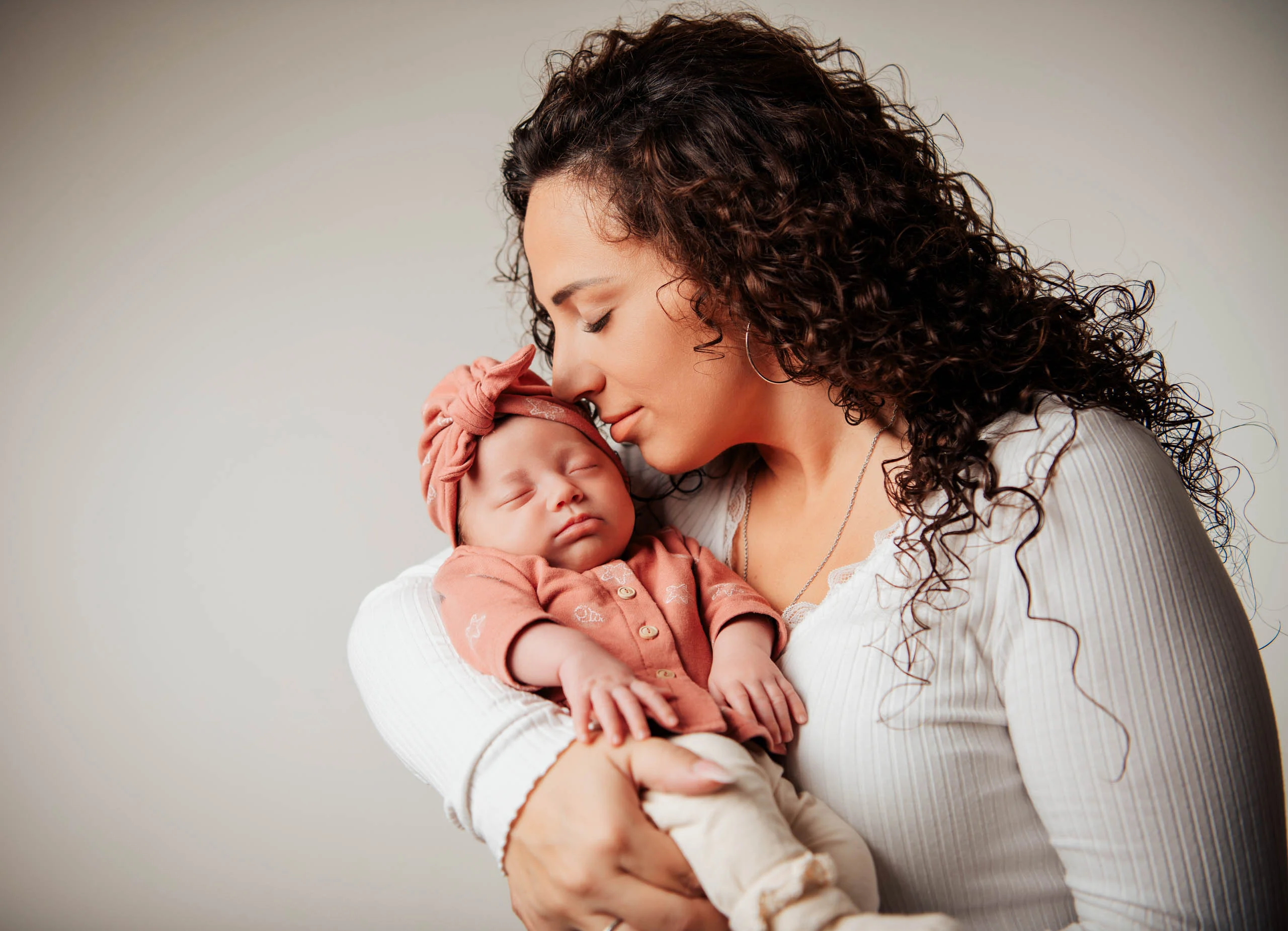 Babyfotos Neugeborene Fotogalerie 069 | Peter Lauritis Photography Frau mit dunklen Locken drückt ihr schlafendes Baby mit rosa Turban an die Wange