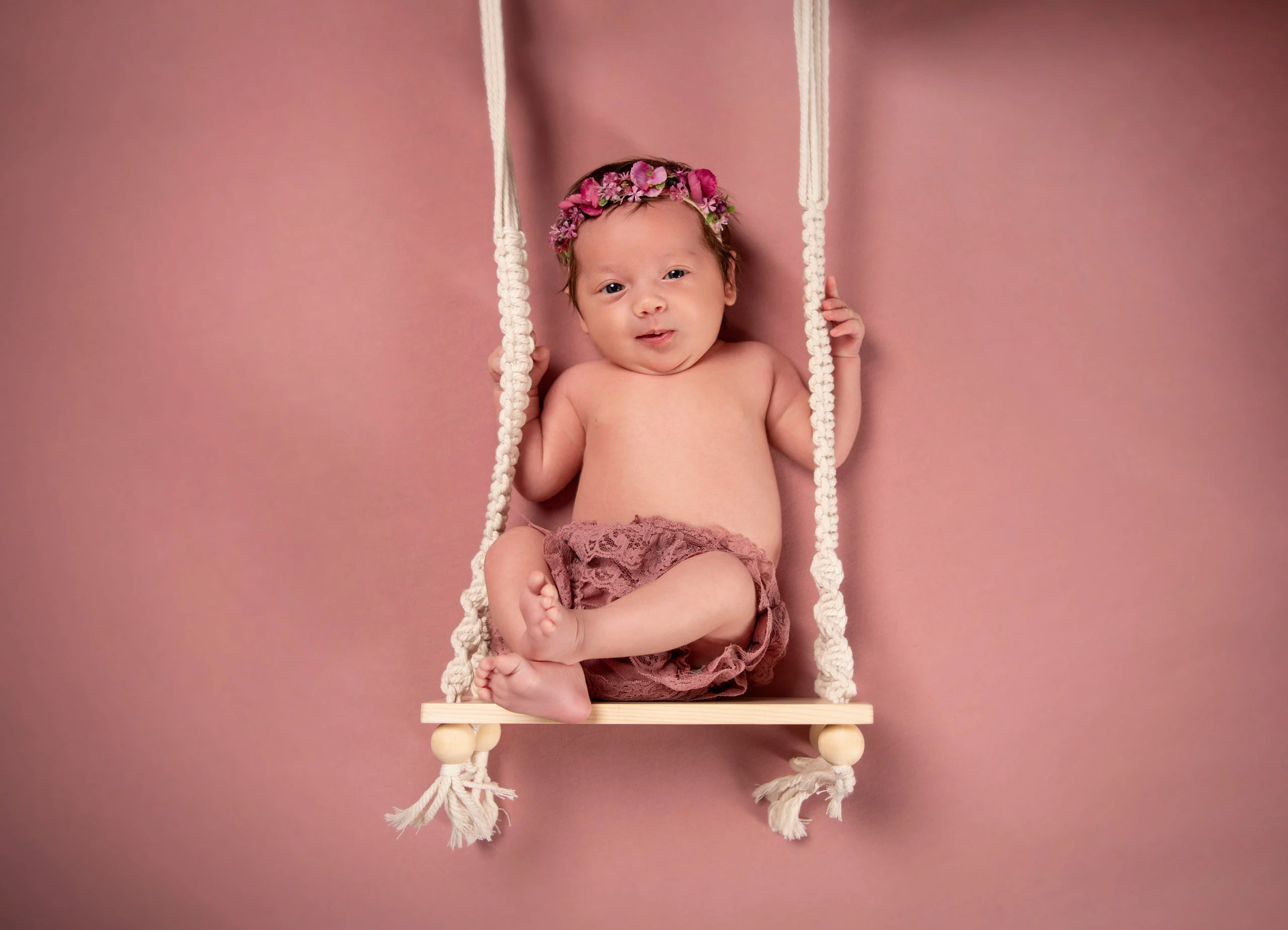 Babyfotos Neugeborene Fotogalerie 070 | Peter Lauritis Photography Baby mit Blumenkranz sitzt auf einer Makramee-Schaukel vor rosa Hintergrund