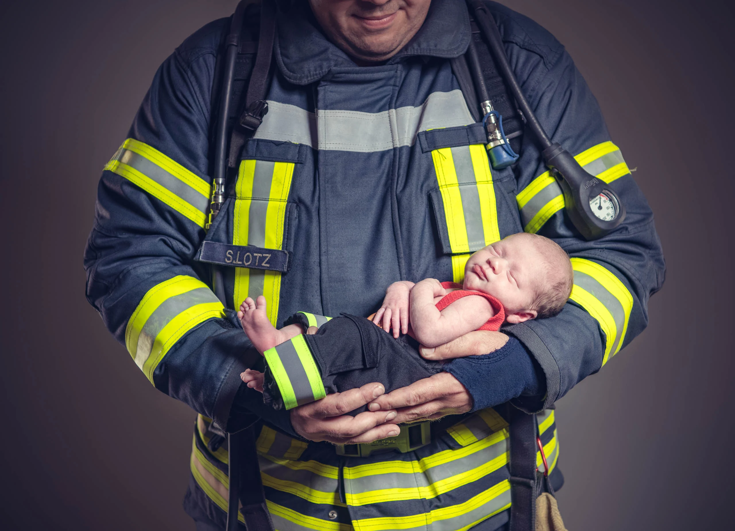 Babyfotos Neugeborene Fotogalerie 101 | Peter Lauritis Photography Feuerwehrmann in Einsatzjacke hält ein schlafendes Neugeborenes