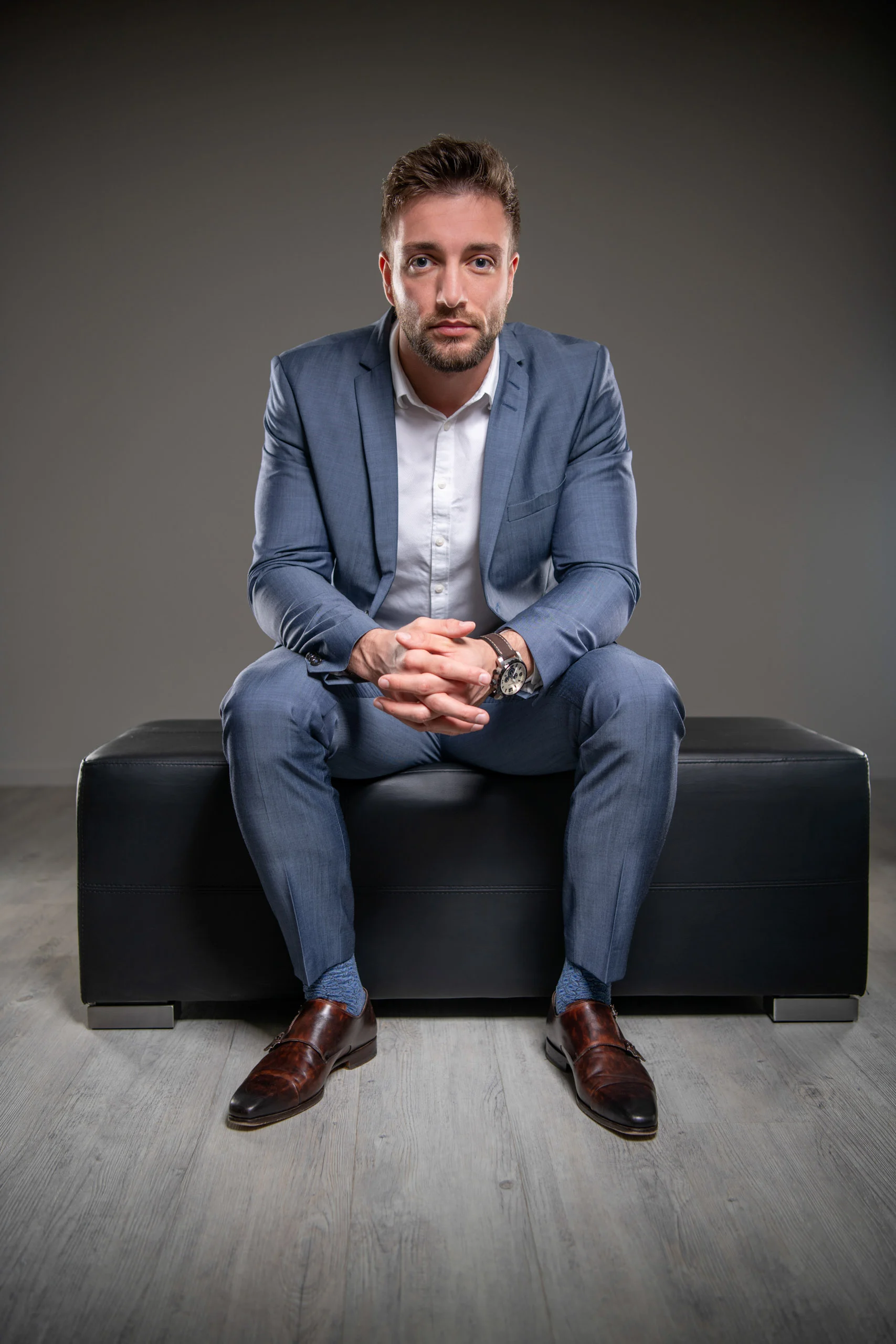 Business Portraits Fotogalerie 021 | Peter Lauritis Photography Mann im blauen Anzug sitzt mit gefalteten Händen auf einem schwarzen Hocker